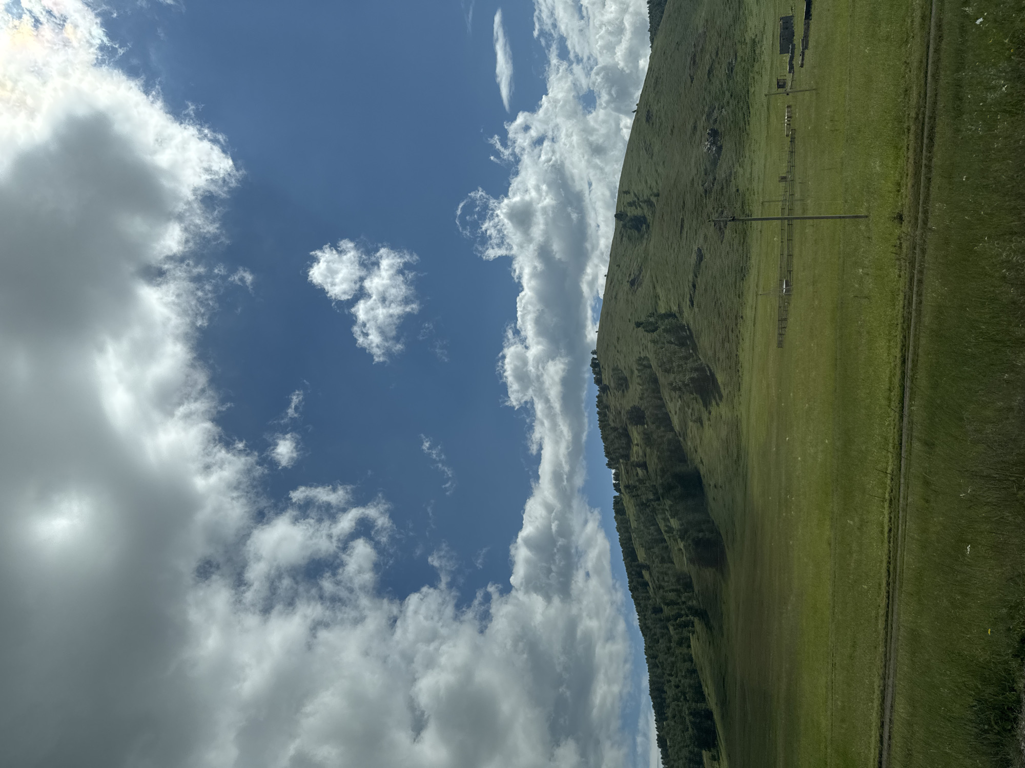 The image depicts a vast green meadow in front of a rolling, tree-covered hill. The sky above is a vibrant blue, dotted with large, fluffy white clouds, with the sun peeking through some of them. In the mid-ground of the meadow, there are several wooden structures, possibly pens or sheds, and a single utility pole. A dirt path or road is visible in the foreground, running horizontally across the frame. The overall impression is one of a rural, natural landscape bathed in sunlight. The location is not specified as being in the city of Uralsk, Russia.
