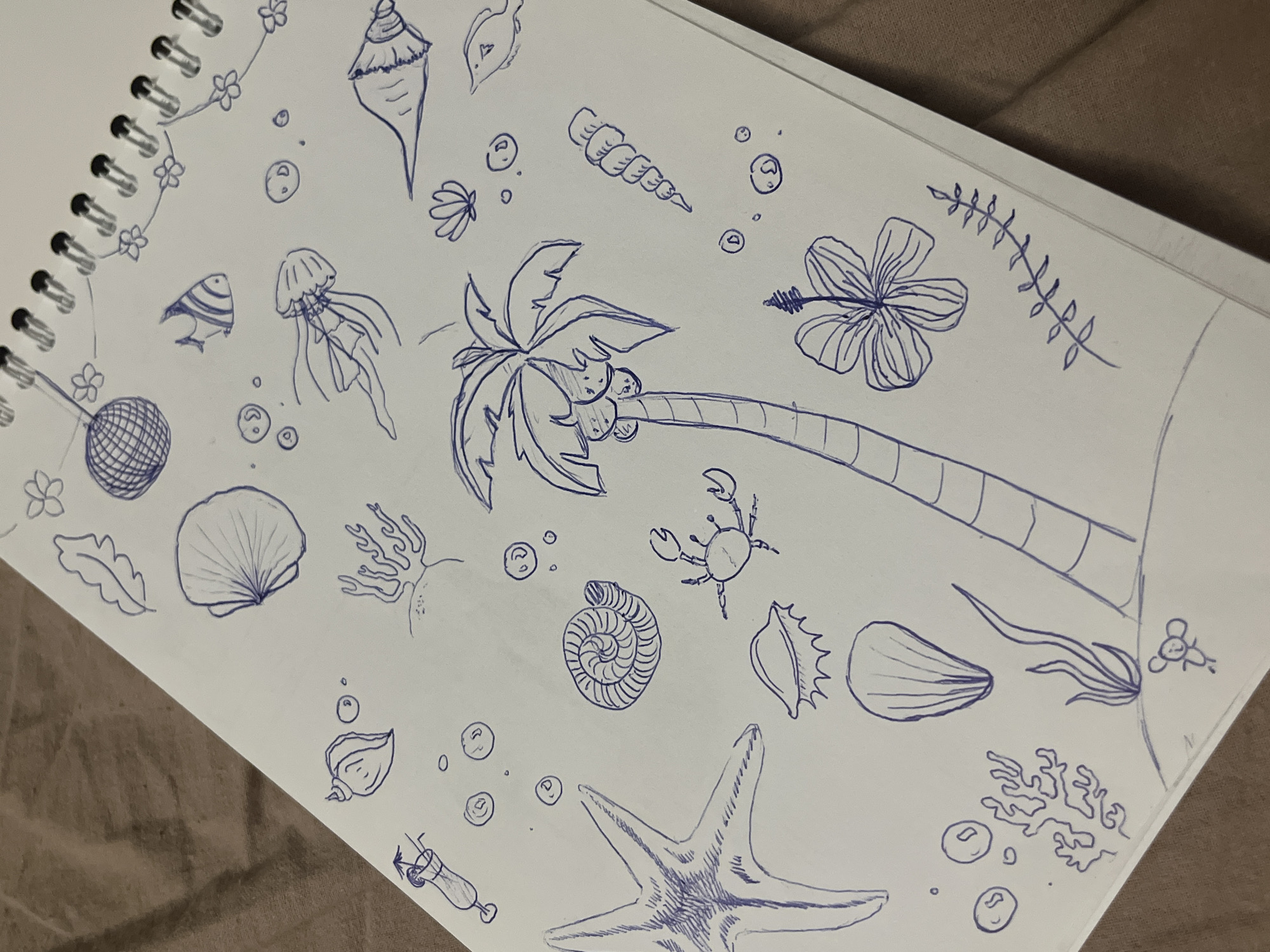 A collection of hand-drawn illustrations featuring marine and tropical themes is displayed on a white, spiral-bound notebook page. The page is angled diagonally across a textured, brown fabric surface.
The drawings, all rendered in blue ink with a line-art style, include a prominent palm tree laden with coconuts, a large hibiscus flower, and a detailed starfish. Surrounding these central elements are various seashells, such as a spiral shell, a conch, a clam shell, and several other distinct types. Marine life is represented by a jellyfish, a small striped fish, a crab, and multiple instances of coral or seaweed. Numerous small bubbles are scattered throughout the composition, suggesting an underwater environment.
Other drawn elements include small flowers resembling plumeria, leafy vines, a hatched spherical object, and a glass containing a drink, possibly a cocktail, positioned in the bottom left. A tiny, simplified drawing that resembles a mouse or small creature is visible in the bottom right corner of the page.
The overall scene captures a creative doodling activity, showcasing a variety of vacation or ocean-inspired motifs. The notebook's spiral binding is visible along the left edge. No text is visible in the image.