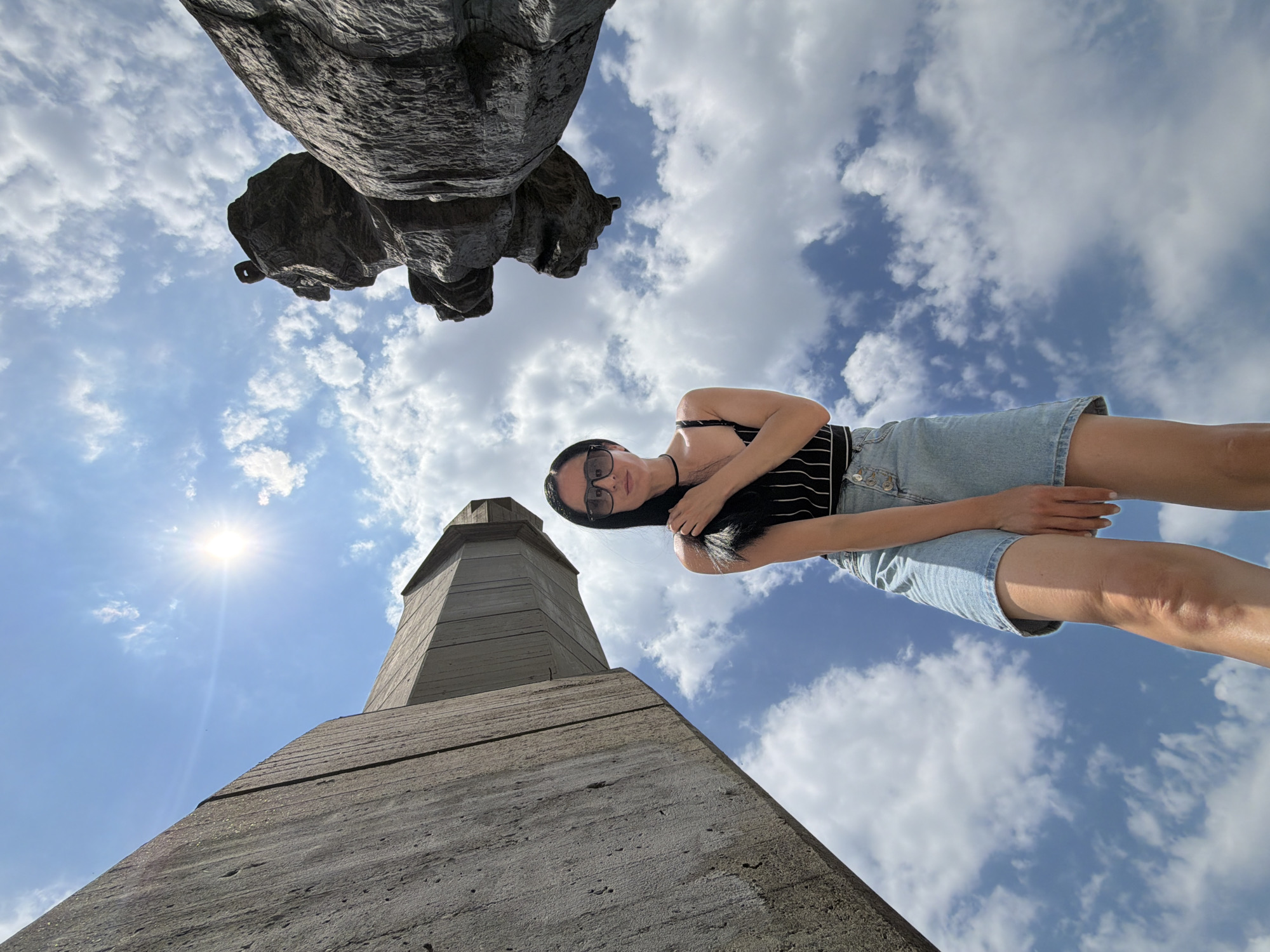 The image captures a low-angle perspective of a woman standing outdoors against a bright, partly cloudy sky. She is wearing a black and white striped crop top and a light-wash denim skirt. Sunglasses are on her face, and she is touching her hair. To her left, a large, imposing concrete structure rises upwards, its rough texture visible. To her right, a portion of a dark, textured sculpture is also prominent. The overall setting appears to be the Pleven Panorama in Pleven, Bulgaria, a historical site featuring monumental structures and sculptures commemorating the Russo-Turkish War.