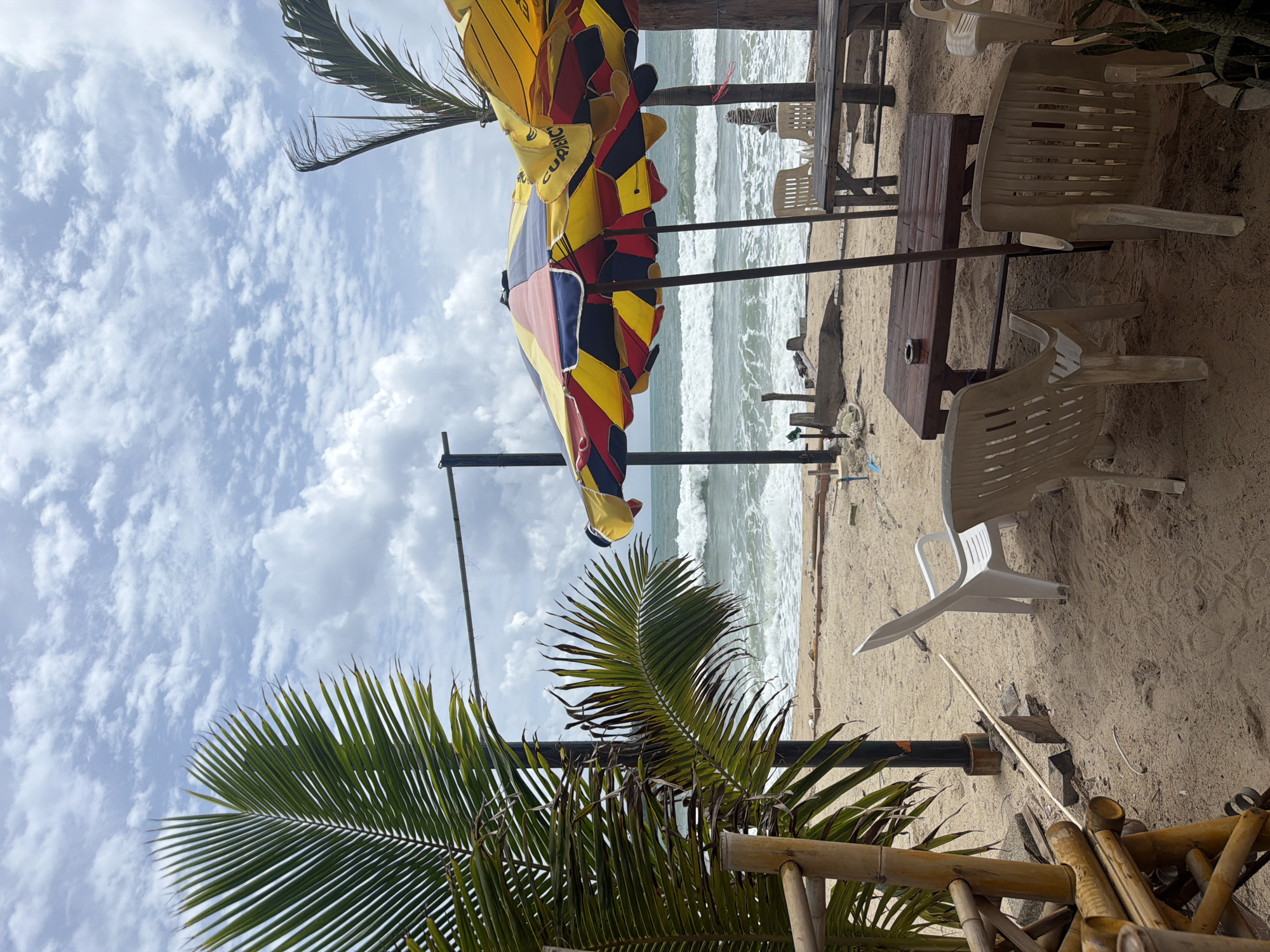 A beach scene in Choeng Thale, Thailand, shows the sandy shore with the ocean and waves in the background under a cloudy sky. In the foreground, a large, colorful umbrella, predominantly yellow, red, and blue, is set up, providing shade. Beneath it, a dark wooden table sits on the sand with several plastic chairs arranged around it. To the left, a portion of a bamboo structure is visible, partially obscuring a palm tree's fronds. Further back, more beach chairs and tables are arranged, some under another umbrella. The overall impression is of a casual outdoor seating area on a tropical beach.