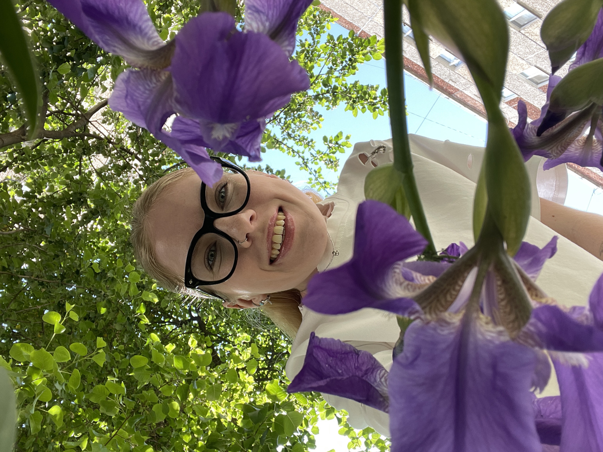A low-angle view captures a woman with fair skin, blonde hair, and black-rimmed glasses, smiling broadly. She has a nose ring and multiple piercings in her ear. She's wearing a white top with what appears to be cut-out details on the shoulder. The subject is surrounded by vibrant purple iris flowers. In the background, green foliage and a blue sky are visible. Additionally, a building can be seen in the distance. While there are no distinct landmarks in the image that suggest Izhevsk, Russia, the style of architecture and surrounding greenery is consistent with a typical urban environment.