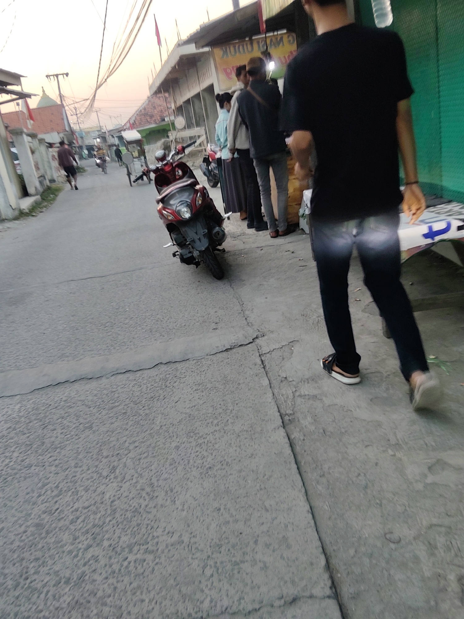 The image depicts a street scene in Mekarmukti, Indonesia, with a focus on a group of people and a motorcycle. In the foreground, a person in a black t-shirt and dark jeans walks away from the camera, wearing sandals. To their left, a red and black scooter is parked on the side of the road. Further down the street, a few other individuals are visible, some walking and others standing. The environment appears to be a commercial area with buildings lining the street and power lines overhead. The lighting suggests it is either early morning or late afternoon. The ground is paved with asphalt and concrete.