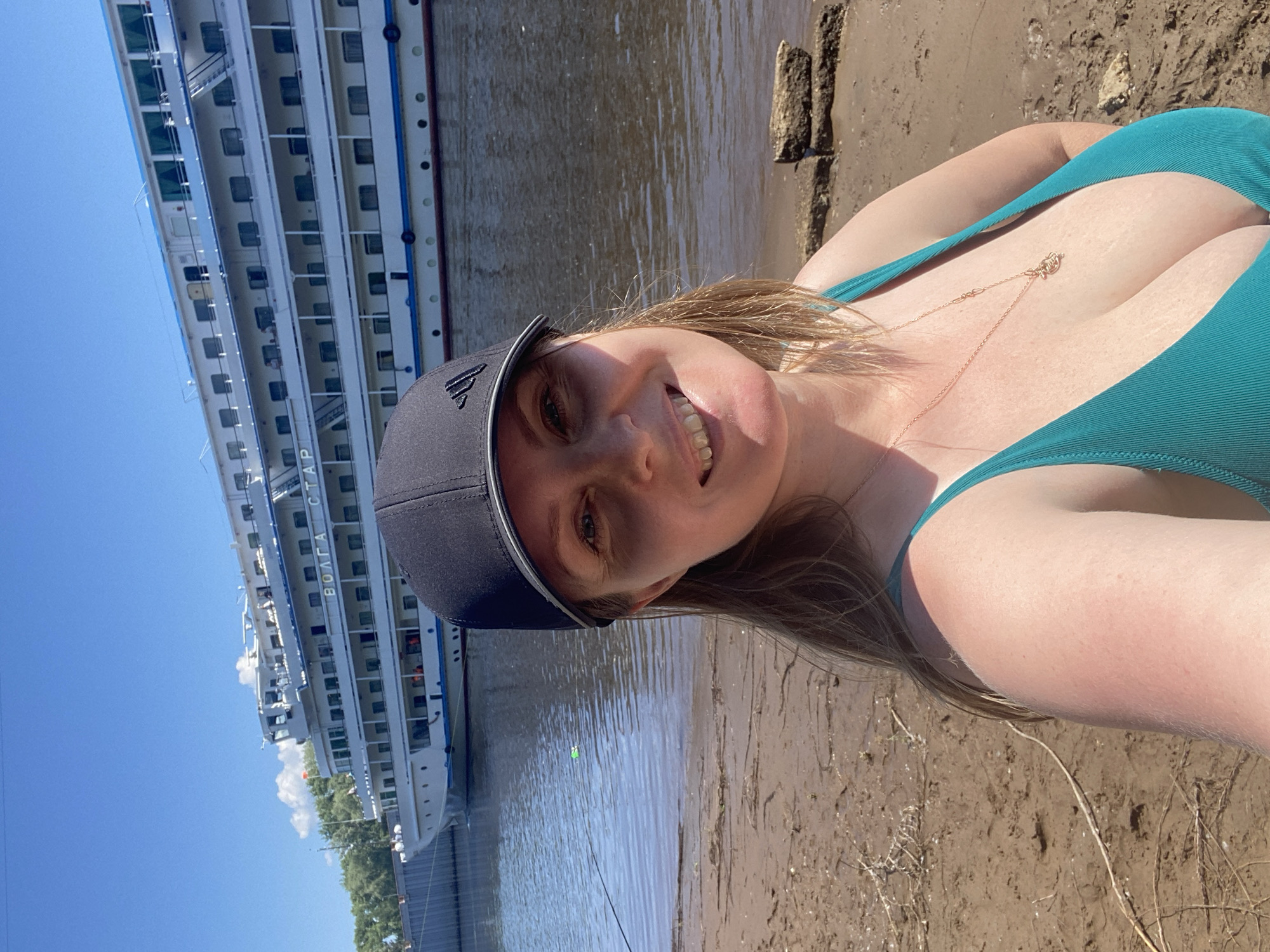 The image is a selfie taken outdoors on a sunny day. In the foreground, a woman with blonde hair, wearing a teal swimsuit and a black baseball cap, smiles at the camera. She is standing on a sandy riverbank. Behind her, a large white cruise ship with multiple decks is docked on the river. The ship has the word "ВОЛГА" visible on its side. The river is calm, and there is a fishing rod with a bobber in the water. The background shows a clear blue sky with a few white clouds and some trees on the distant shore. The setting is consistent with the Kama River embankment in the city of Chaikovsky, Russia, known for its river port and cruise ship traffic.