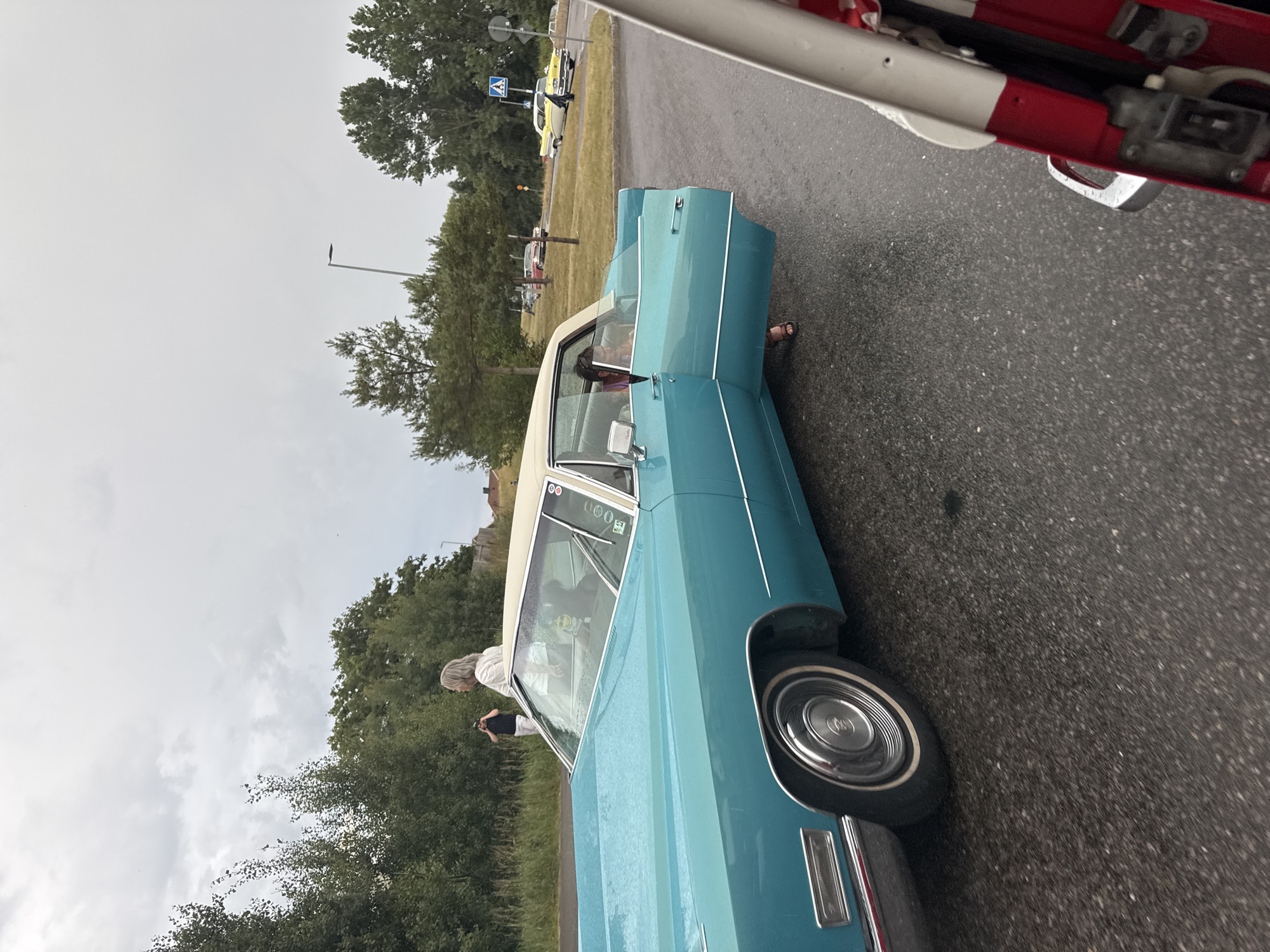 The image depicts a cloudy day in Falkenberg, Sweden, likely during an event or gathering related to vintage cars. The foreground features a turquoise Cadillac with its driver's side door open. A person with dark hair, wearing a pink shirt, is seated in the driver's seat, looking towards the front. To the left of the Cadillac, a woman with blonde hair, wearing a white top, is standing and holding what appears to be a camera. Further in the background, another person in a dark shirt and white pants is visible. Several other classic cars, including a yellow and a red one, are parked or driving in the distance on a wet asphalt road. Trees and vegetation line the background, suggesting a location near a park or a residential area. The overall scene captures a moment during an outdoor car exhibition or cruise.