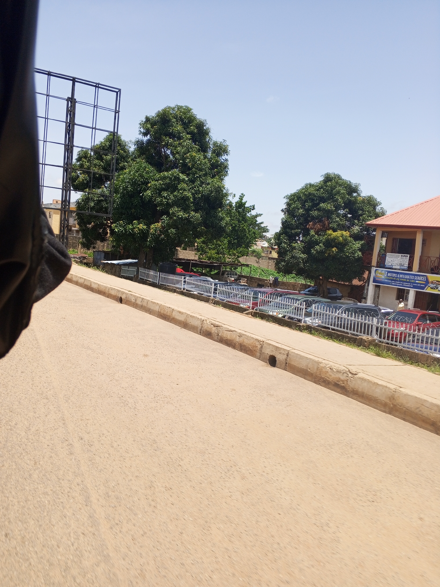 A low-angle, full shot captures a street scene in Kabong, Nigeria. In the foreground, a beige asphalt road curves to the left, bordered by a concrete curb. Beyond the curb, a white metal fence encloses a lot filled with cars, suggesting a car dealership or parking area. Several vehicles are visible, including what appears to be a red SUV. A multi-story building with a red-tiled roof stands to the right, featuring signs that are partially visible. Trees with lush green foliage dot the background, with one particularly large tree dominating the center of the frame. A tall metal structure, possibly a billboard frame, is visible to the left, partially obscured by a dark, out-of-focus object in the extreme left foreground, which might be part of the vehicle from which the photo was taken. The sky is clear and light blue, indicating a sunny day.