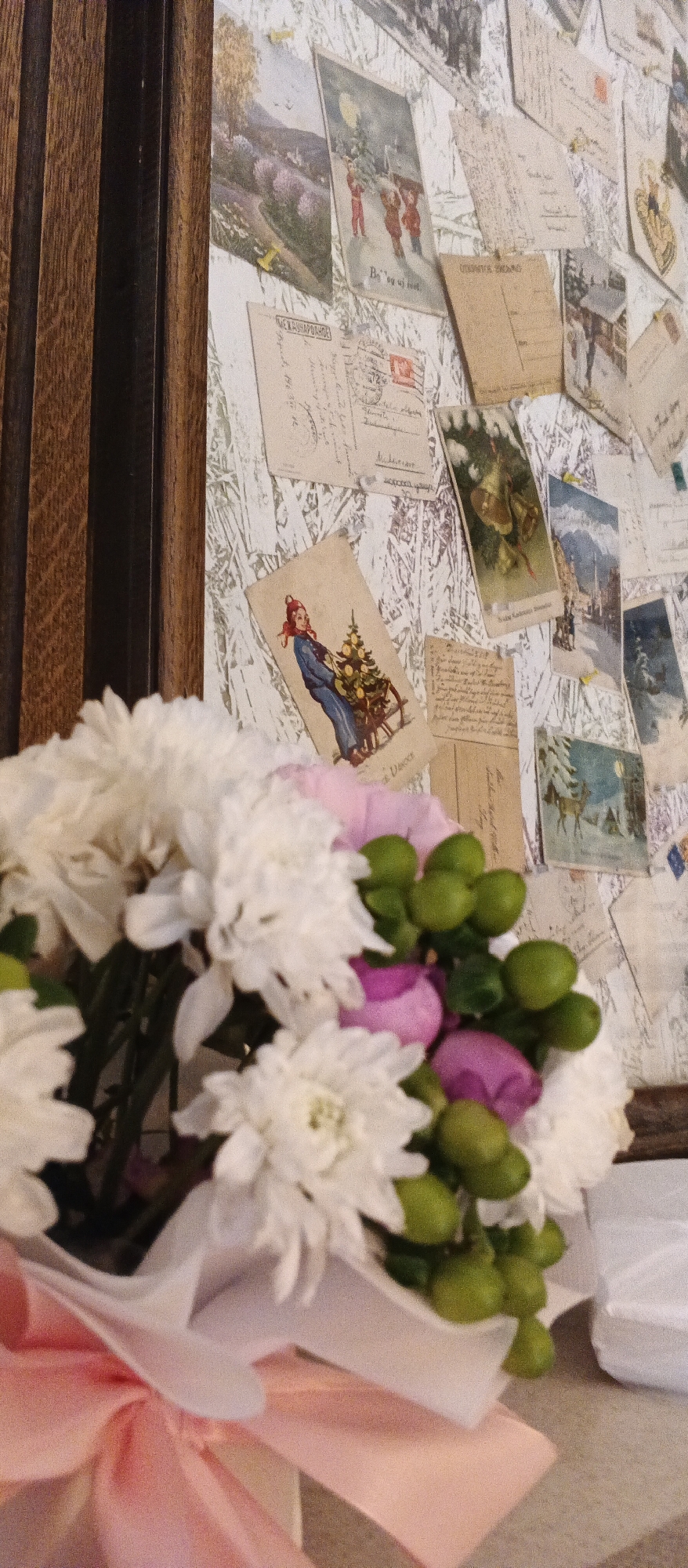 The image shows a bouquet of white chrysanthemums and pink roses with green berries, tied with a pink ribbon, in the foreground. In the background, a wall is decorated with old postcards, some of which depict winter scenes and people. The wall appears to be a backdrop in a cafe or shop. The room has wooden paneling on the left side. The overall impression is a cozy, perhaps vintage, interior. The image was taken in Berehove, Ukraine.
