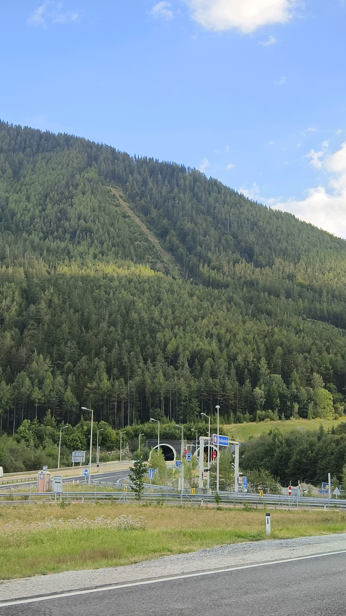 The image depicts the motorway leading to a tunnel entrance near Schottwien, Austria. The scene is dominated by a dense, green forest covering a steep mountain slope that rises in the background. A diagonal, lighter-colored scar runs up the mountainside, suggesting a cleared area or a path through the trees. In the foreground, a highway with multiple lanes is visible, bordered by grassy verges and some vegetation. Several streetlights line the road leading up to the tunnel. The tunnel entrance itself is a dark, arched opening, and above it, there are gantries with traffic signals and signs. The sky is blue with scattered white clouds. There are no people or vehicles clearly visible in the frame, giving the impression of an empty, scenic view of infrastructure integrated into a natural landscape.