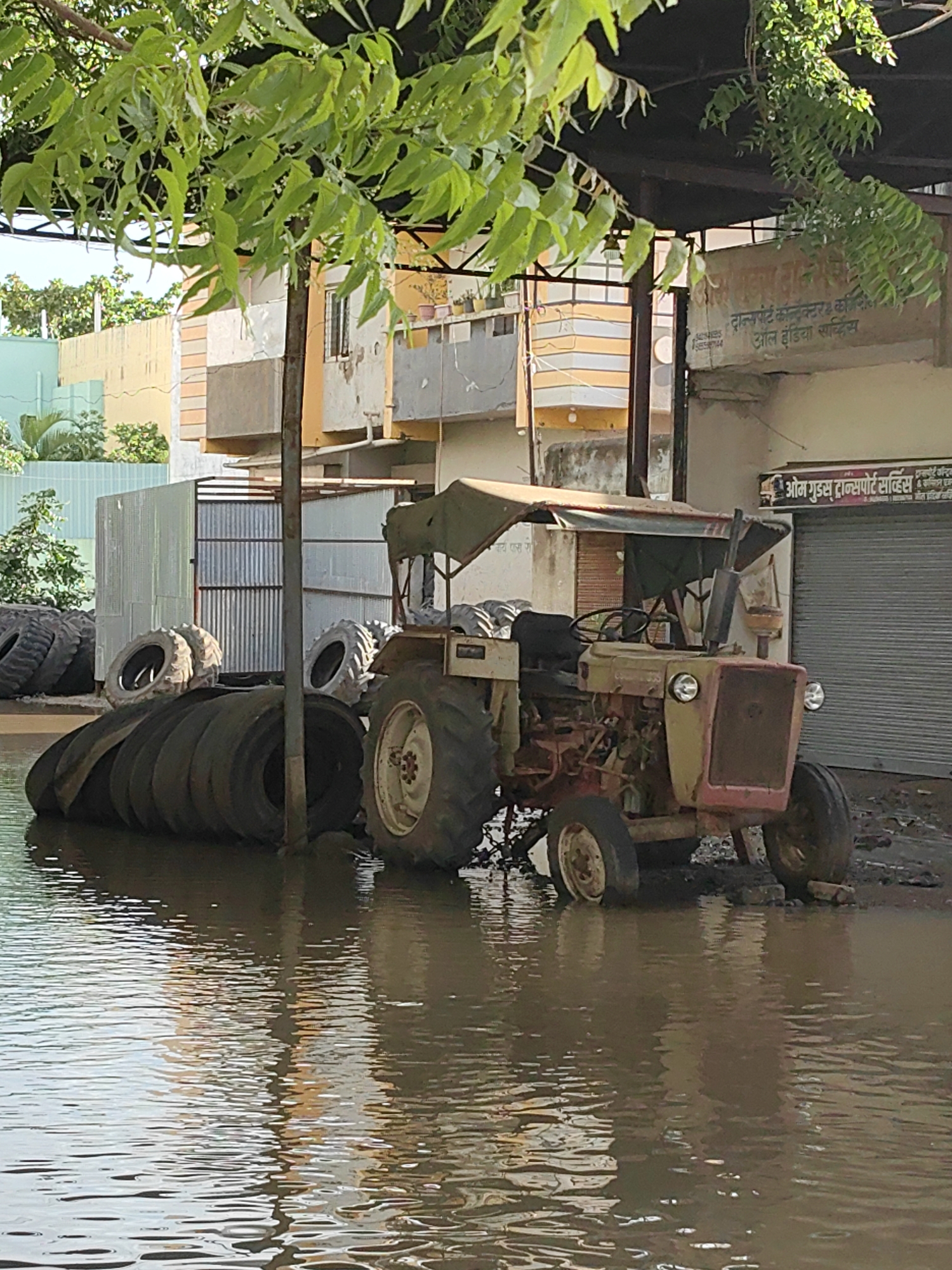 The image depicts a flooded scene in the city of Malkapur, India, likely an industrial or commercial area given the presence of a tractor, stacked tires, and buildings with signage. A light-colored tractor, with a red front grille and a canvas canopy, is partially submerged in murky floodwaters. Several large tires, some stacked and others leaning, are also in the water, some appearing to be used tractor or vehicle tires. In the background, a building with beige and orange striped sections is visible, along with corrugated metal structures. One building has a sign that reads "ओम गुड्स ट्रांसपोर्ट सर्विस" (Om Goods Transport Service), suggesting a business operation. The ground is uneven and appears to be a mixture of mud and water. Green foliage from a tree overhangs the scene from the top of the image, casting shadows. The overall impression is one of disarray and the impact of heavy rainfall or flooding.