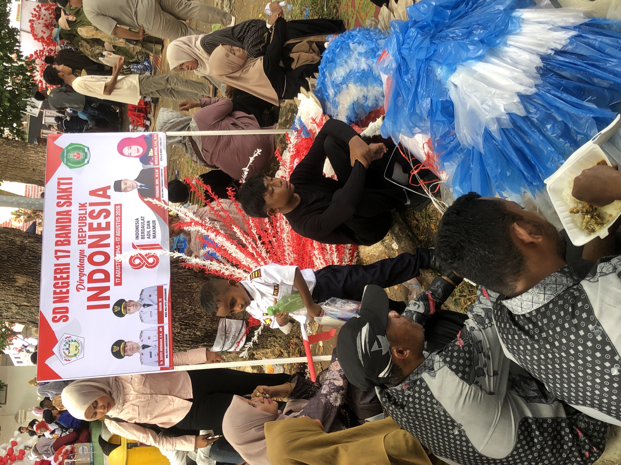 The image depicts a gathering outdoors, likely a public event, in Lhokseumawe, Indonesia. In the foreground, several individuals are seated on the ground, some of whom are eating. One man in the lower right corner is eating from a white Styrofoam container filled with rice and what appears to be some sort of stew or curry. He is wearing a patterned black and white long-sleeved shirt. To his left, another man in a similar shirt is partially visible. A young boy in a white pilot uniform with a green bottle in his hand stands to the left of center, looking towards the right. He is holding a clear plastic bag.
Behind the seated individuals, a large banner is prominently displayed, attached to a tree. The banner reads "SD NEGERI 17 BANDA SAKTI" and "Dirgahayu REPUBLIK INDONESIA". It features images of men in official attire, likely politicians or local leaders, and text indicating "17 AGUSTUS 1945 - 17 AGUSTUS 2026". Red and white decorative streamers are hung around the banner and trees.
In the background, a crowd of people are visible, including women wearing hijabs and men in various casual and official attire. The environment appears to be a park or open area, with trees and foliage present. Several yellow and blue plastic bags are scattered around the scene, contributing to the cluttered appearance of the ground. The overall impression is of a community celebration or event.