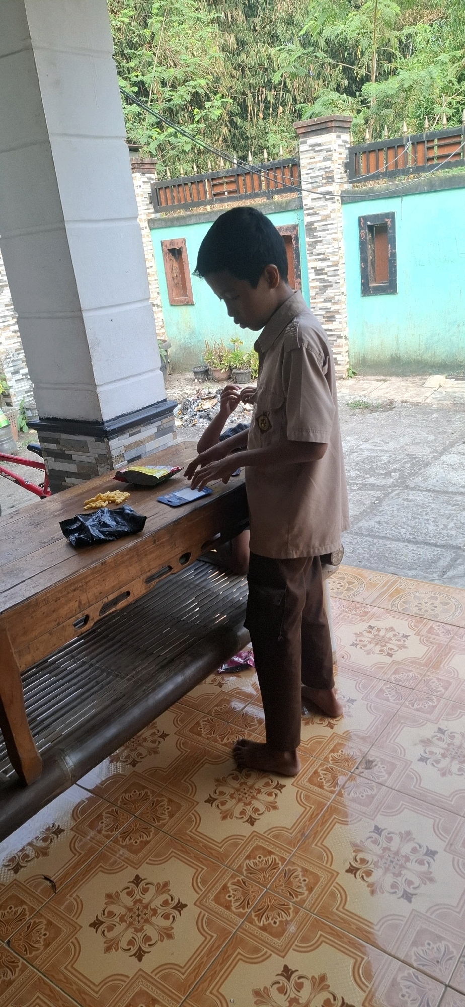 In Cibinong Hilir, Indonesia, a young boy wearing a tan uniform shirt and brown pants stands barefoot on a tiled patio. He is looking down at a wooden table where a smartphone, a black plastic bag, and a bag of snacks are placed. The boy's hands are near the table, suggesting he might be interacting with the phone or the snacks. Behind him, a turquoise wall with wooden-framed windows is visible, and beyond that, lush green foliage. The patio's floor is covered in patterned orange and cream tiles, some of which appear wet. A portion of a white pillar and a bamboo bench are also visible in the scene.