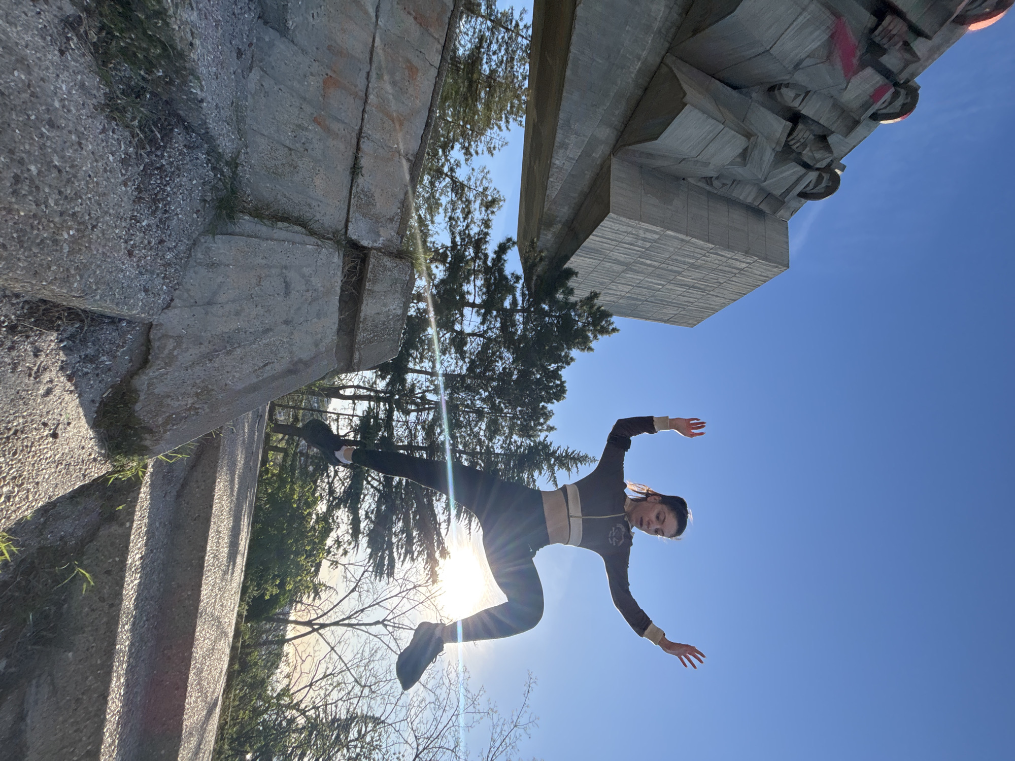 The image shows a young woman wearing a dark athletic outfit with light-colored accents, posed mid-air in a dynamic, acrobatic pose against a bright blue sky. The background includes concrete structures of the Monument of the Bulgarian-Soviet Friendship in Varna, Bulgaria, and trees. The sun is positioned behind the woman, creating a backlit effect. The image is oriented as if the monument and the surrounding landscape are sideways, creating an illusion of the woman defying gravity. The monument features geometric concrete blocks, while the surrounding area shows typical urban landscaping with trees.