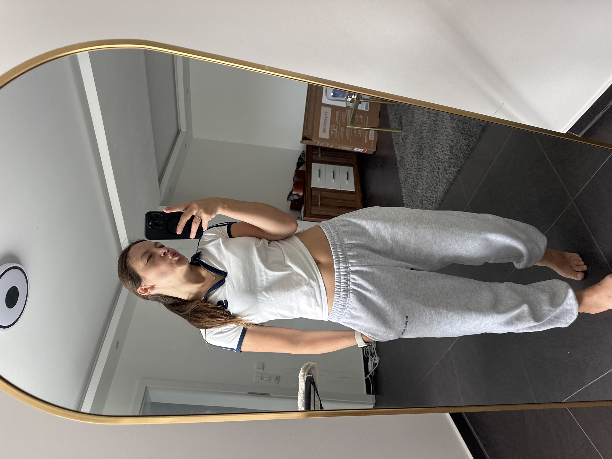 The media file is a full-body mirror selfie taken by a young woman. She is wearing a white t-shirt with navy blue trim and light grey sweatpants. The woman is standing in what appears to be a modern, well-lit room with dark tiled flooring and a grey textured rug. In the background, reflected in the mirror, is a dresser with white drawers, a cardboard box labeled "CLARION FHD 20", and part of a guitar. The woman is puckering her lips and holding a black smartphone in her right hand, taking the photo. She has long brown hair tied back in a ponytail. A white circular object with a black center is visible on the wall to her left. The overall impression is casual and informal, possibly taken at home. There is no visible text besides what is on the cardboard box. The time of day and weather are not discernible from the image. The location context is Kutenholz, Germany.