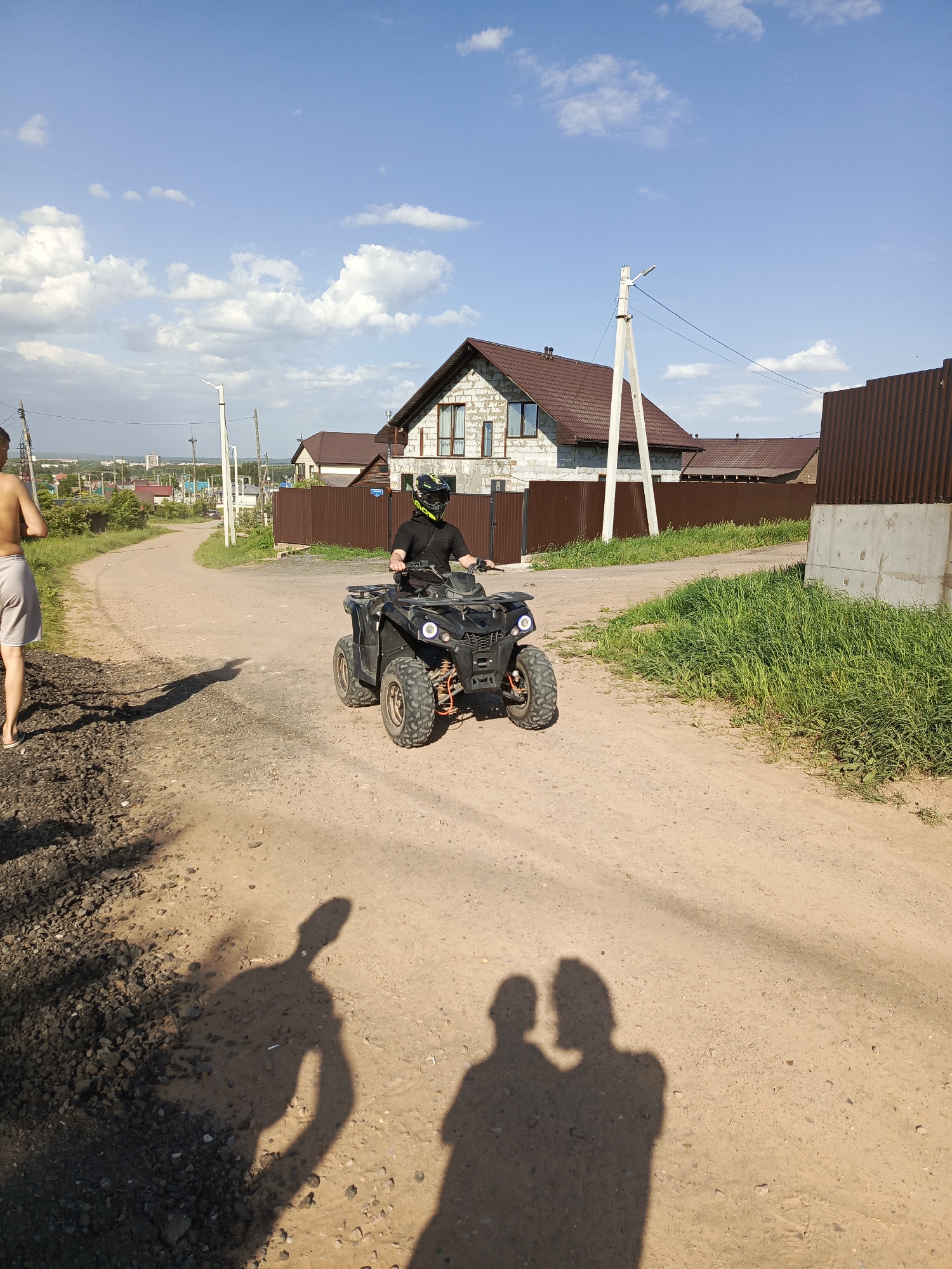 The image shows a person driving a black ATV on a dirt road. The driver is wearing a black shirt and a helmet. To the left of the ATV, a shirtless person is walking. In the background, there are houses, a fence, grass, and utility poles. The sky is blue with scattered clouds. The location is in Chaikovsky, Russia. Shadows of people taking the photo can be seen in the foreground.