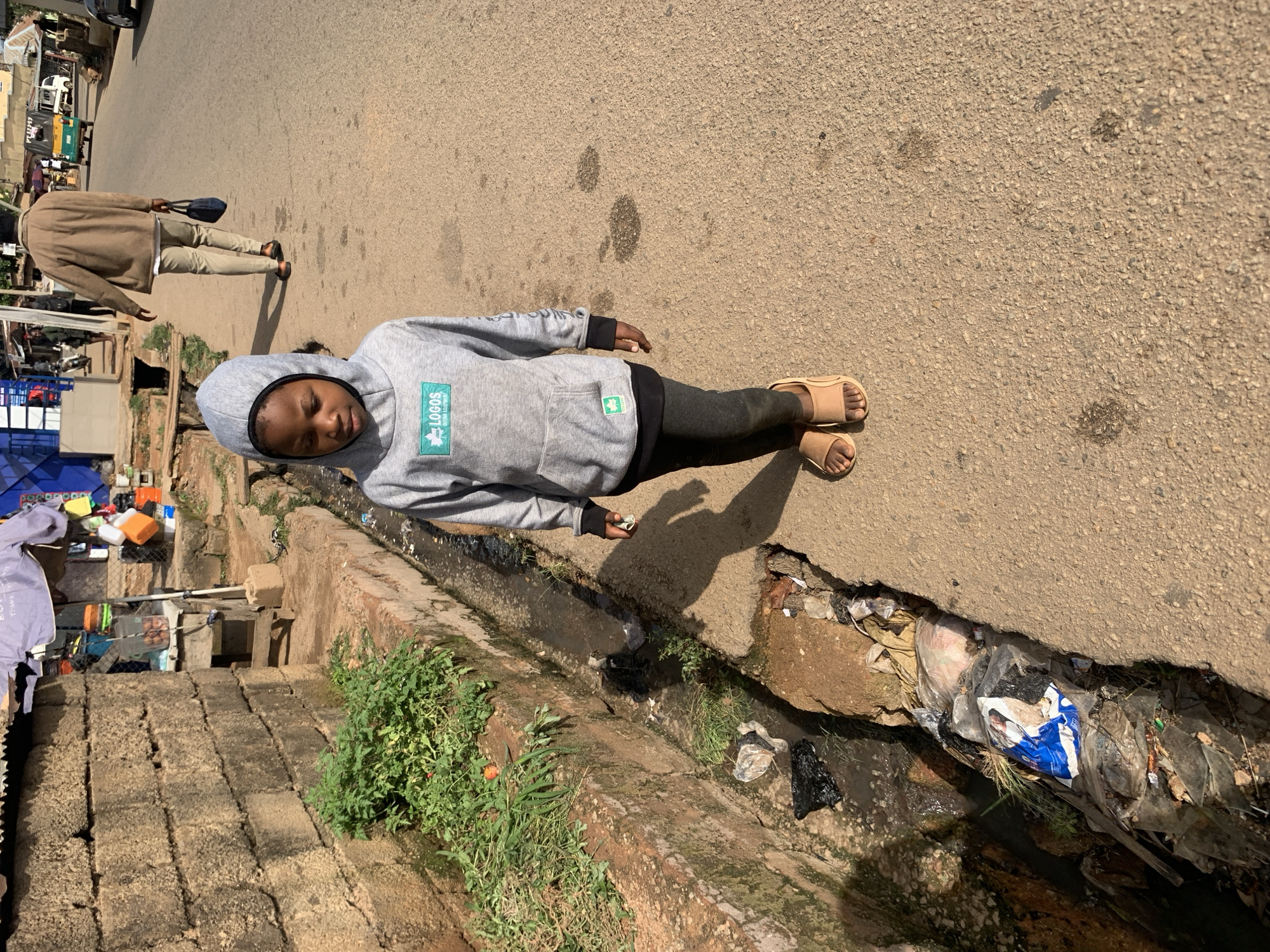 The image depicts a street scene in Gura Top, Nigeria. A young child wearing a grey hooded sweatshirt and dark leggings stands on a paved road. The child has dark hair and is looking downwards. To the left of the child is a drainage ditch filled with water and debris, including plastic bags and what appear to be discarded food wrappers. Green vegetation grows along the edge of the ditch. In the background, a man in a brown jacket walks away from the camera, carrying a dark bag. Further in the background, a white truck and a yellow and green motorized rickshaw are visible on the road. The buildings on the left are constructed of rough stone blocks. The overall scene suggests a common urban environment with evidence of both commerce and informal settlements.