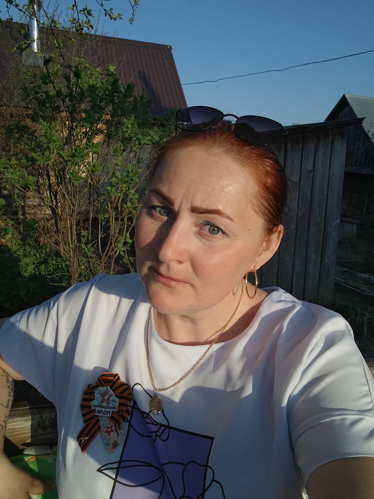 A woman with red hair and green eyes is the primary subject of the image. She is wearing a white t-shirt with a floral graphic and an orange-and-black ribbon pinned on her chest. Sunglasses rest on top of her head, and she is wearing gold hoop earrings and a gold necklace with a pendant. She is facing the camera with a neutral expression.
The background consists of foliage, a wooden fence, and two buildings. The location is Фоки, Russia, indicated by the Cyrillic script on the ribbon. The scene is lit by sunlight, suggesting it is outdoors during the day.