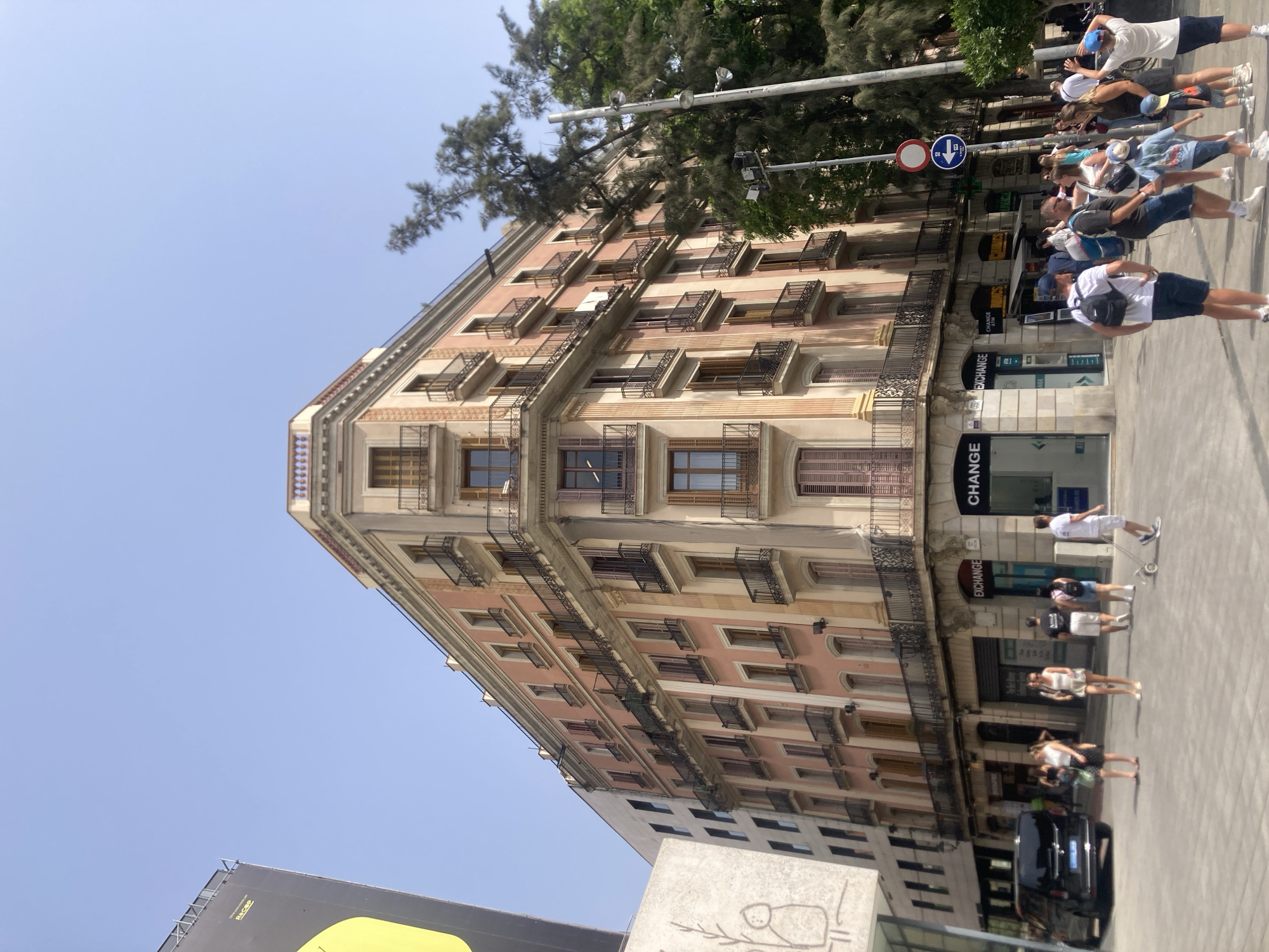 This is a street-level, medium-angle, outdoor, daytime photograph taken in Barcelona, Spain. The image is dominated by a large, multi-story building with a pinkish-orange facade and light brown trim. The building features numerous balconies with black wrought-iron railings. Several windows on the upper floors are visible, some with wooden shutters. The building appears to be on a corner, with its facade extending along two streets.
In the foreground, there is a paved walkway or plaza with several people walking and standing. A black minivan is parked on the street to the left. Several businesses with signage are visible at the ground level of the building, including two "EXCHANGE" offices. A large, modern billboard with a yellow graphic is partially visible on the far left, behind a contrasting light-colored building with a simple line drawing on its exterior. A tall tree with dark green foliage is situated to the right of the main building, obscuring part of its upper floors. Two streetlights are mounted on a pole to the right of the tree. A circular blue and white directional sign is visible above the street level businesses. The sky is clear and blue.
