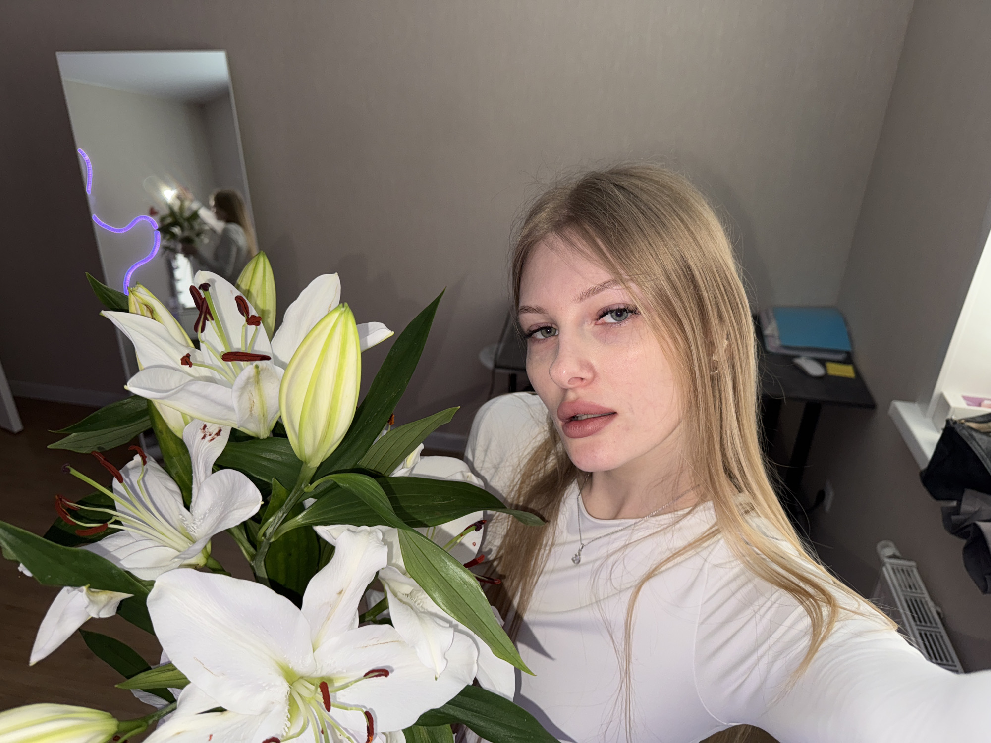 A young woman with fair skin and blonde hair stands indoors, holding a bouquet of white lilies with green foliage. The flowers are in the foreground, partially obscuring her torso. She is wearing a white, long-sleeved top and a delicate silver necklace. Her gaze is directed towards the camera.
Behind her, a mirror reflects the scene, showing a partial view of the room and the woman herself holding the flowers. In the background, a desk with papers and other objects is visible, as well as a window providing natural light. A decorative light strip is seen along the edge of the mirror.
The setting appears to be a room within a residential dwelling. There is no information in the image to suggest a specific location in the city of Perm, Russia.