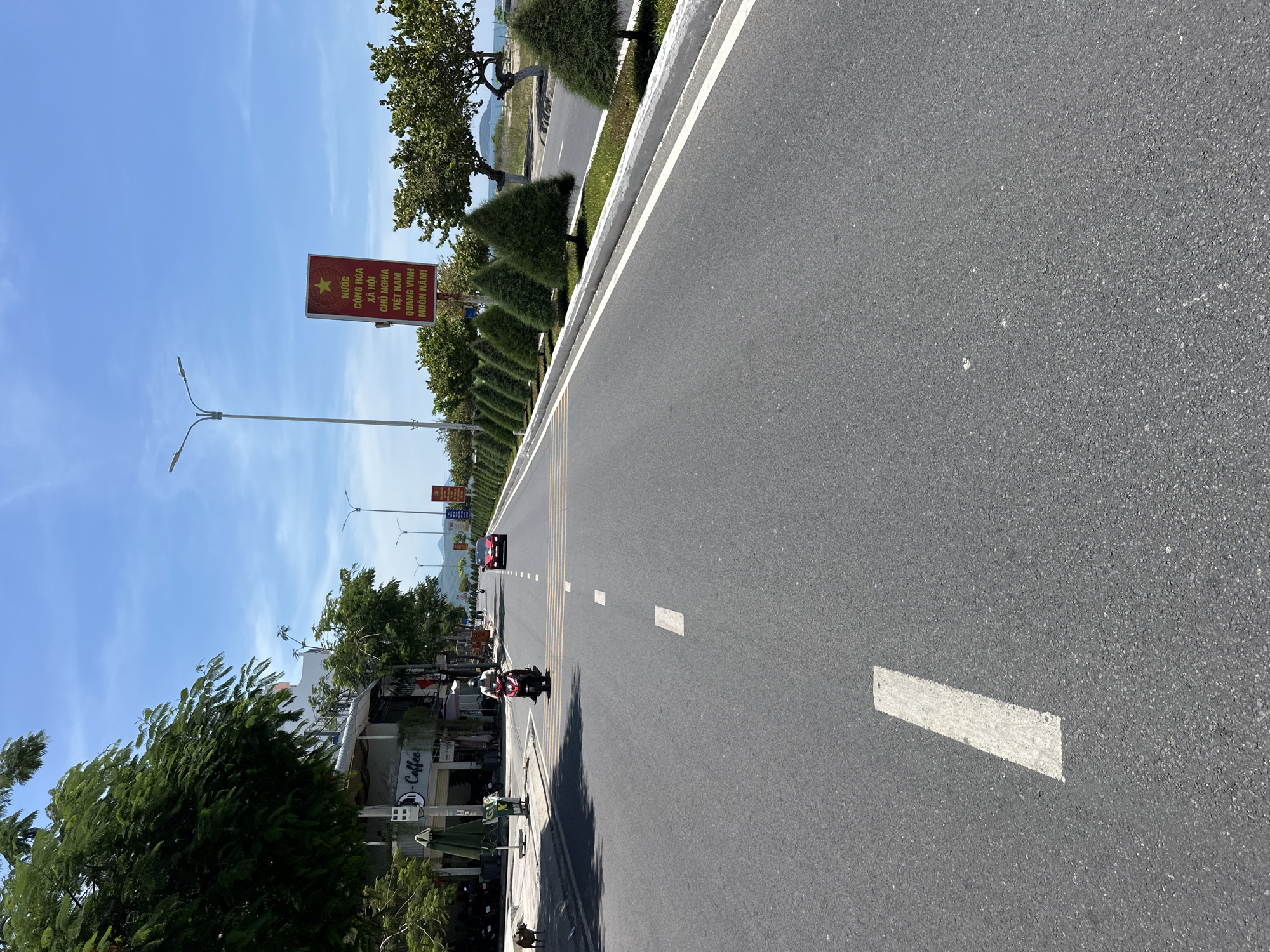 The image depicts a straight, paved road in Bac Nha Trang Ward, Vietnam, under a clear blue sky. On the left side of the road, there are buildings with awnings and signage, including one that reads "- Coffee". A scooter with a rider wearing a helmet is in motion on the road, approaching a pedestrian crossing marked with white stripes. A red car is visible further down the road. To the right of the road, there is a row of small, cone-shaped trees planted along a white concrete curb. Beyond the trees, a body of water and distant hills are visible. A tall lamppost stands near the right edge of the road, next to a red sign with Vietnamese text and a yellow star. A stray dog is present on the sidewalk on the left side of the road.
