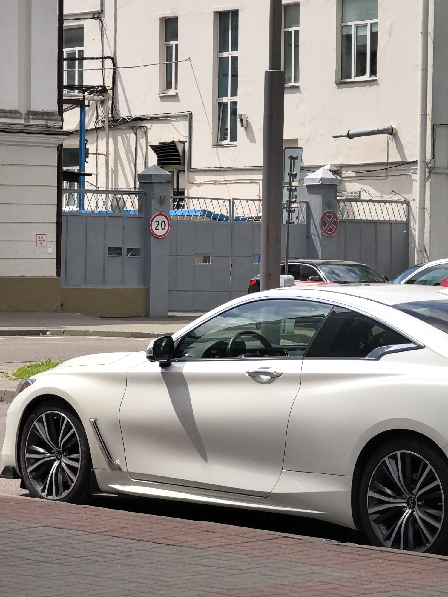 An overhead, eye-level shot shows a white coupe car parked on a city street. The car's sleek design is highlighted by its low profile and sporty wheels. The car is positioned on a brick-paved road, with a sidewalk and a grassy area to its left. In the background, a grey building with multiple windows stands behind a metal fence topped with barbed wire. A "20" speed limit sign is visible on the fence, as well as other signs and pipes attached to the building. The scene appears to be on a bright, sunny day.