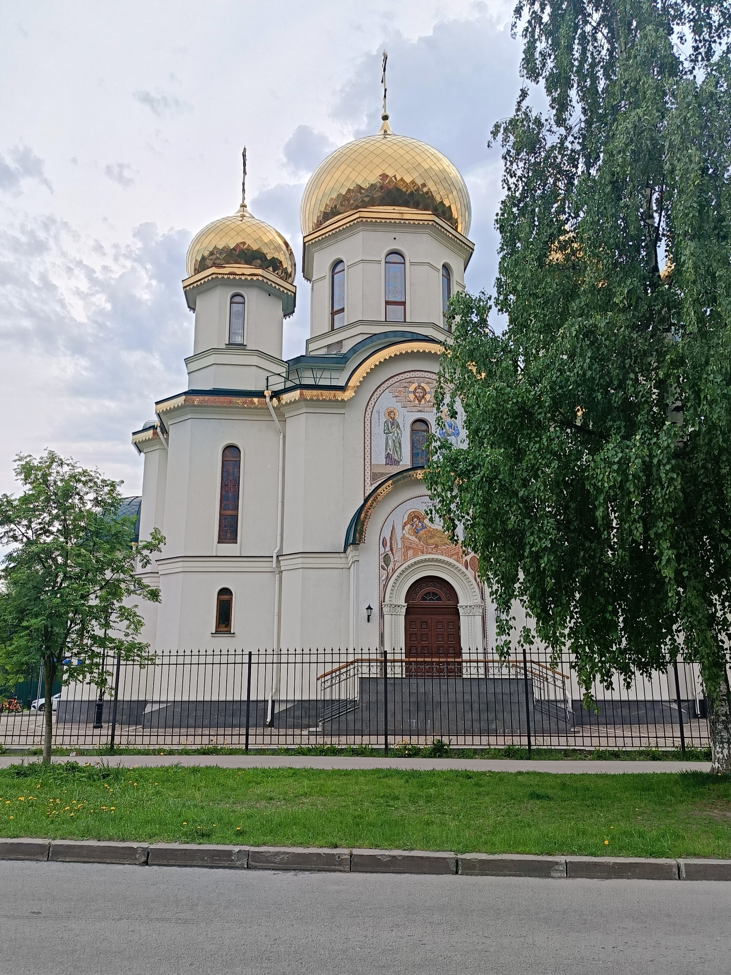 The image depicts a white Orthodox church with golden domes in Saint Petersburg, Russia. The church is surrounded by a black metal fence. Green trees partially obscure the view of the building. A paved road and a grassy area with dandelions are in the foreground. The sky is overcast. No people are visible in the scene.