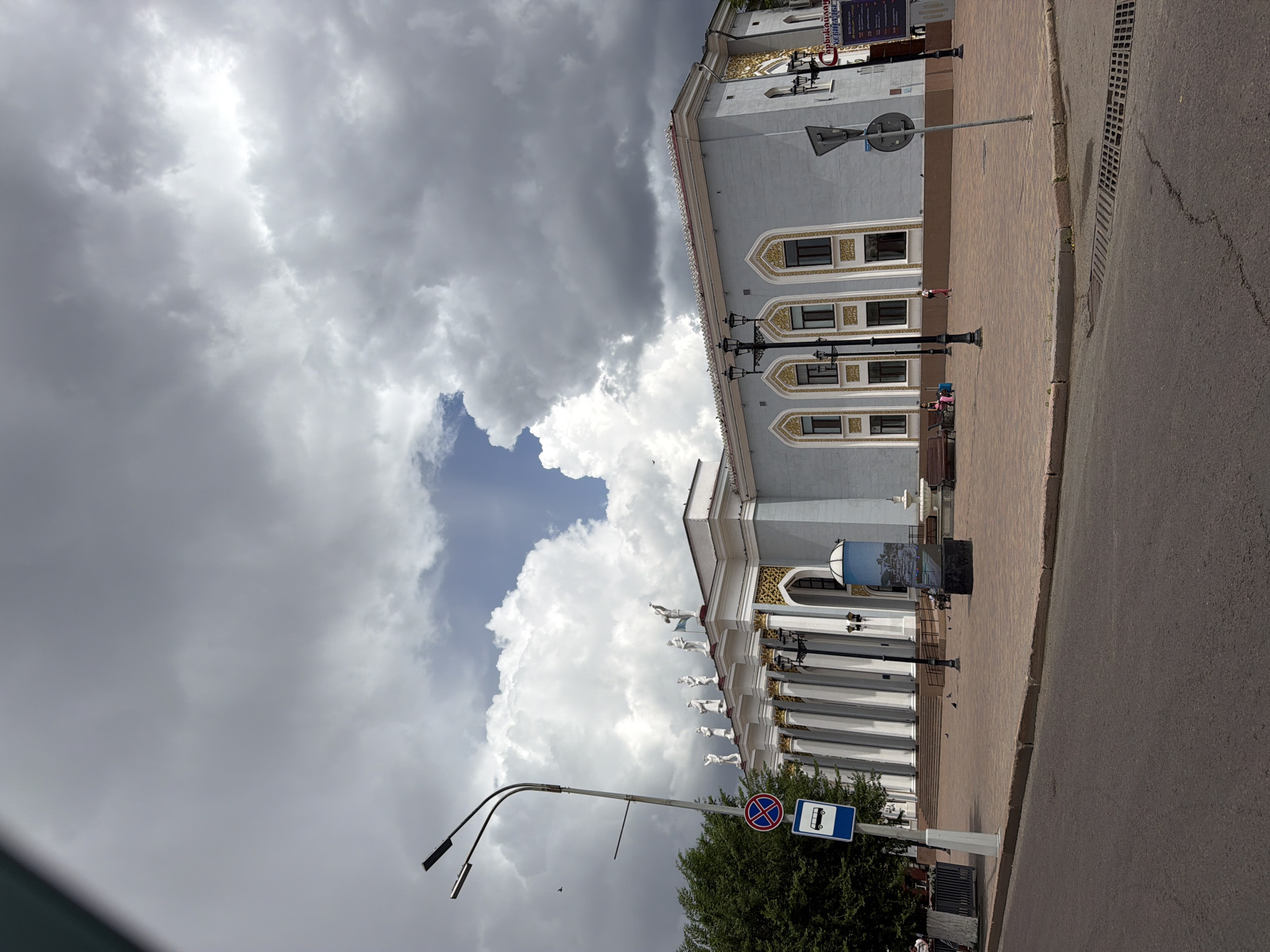 The image depicts the city administration building in Qarağandı, Kazakhstan, under a dramatic, cloudy sky. The building features a classical facade with a series of white columns supporting an ornate roofline, adorned with statues of figures. Several windows with decorative gold framing punctuate the pale blue exterior walls. In front of the building, a paved plaza is visible, with a street lamp and a bus stop sign. Pigeons are scattered across the plaza. A few people are present, including a person on a bench and another walking in the distance. To the left of the frame, a large tree adds greenery to the scene, contrasting with the architecture and the stormy sky. The foreground shows a section of asphalt road.