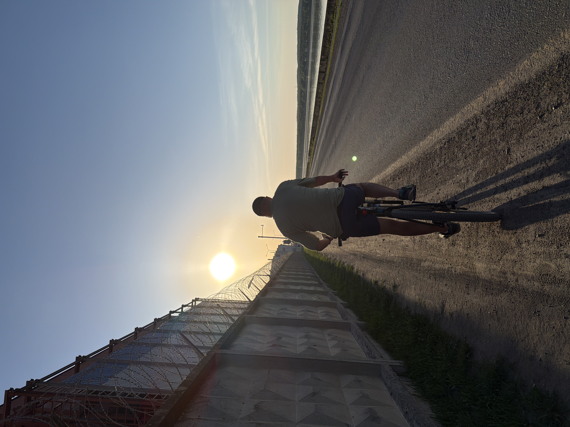A man rides a bicycle on a road in Chaikovsky, Russia. The photograph is taken at a low angle, with the sun setting or rising on the left side of the frame. To the left of the road is a concrete wall topped with barbed wire. The man is silhouetted against the light, but he appears to be wearing shorts and a short-sleeved shirt. The road stretches into the distance. In the background, there is a body of water and some vegetation.