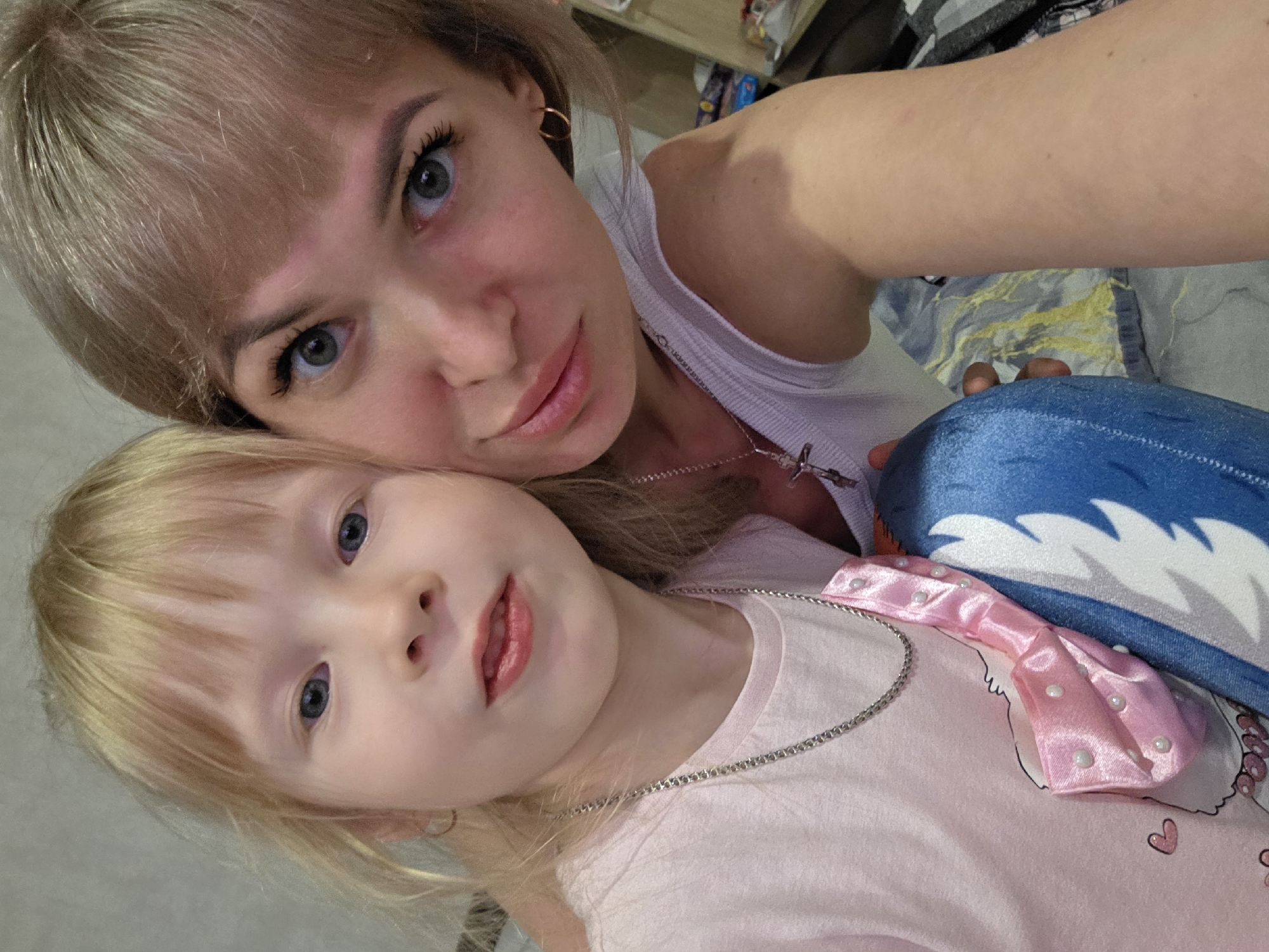 This close-up indoor portrait features an adult female and a young child, likely a mother and daughter, captured in a selfie-style shot. Both subjects have blonde hair and light-colored eyes, with bangs.
The adult is positioned in the upper part of the frame, with her head slightly tilted. She has a serene expression, looking directly at the camera. She wears a white ribbed tank top and a silver chain necklace with a cross pendant. A small gold hoop earring is visible in her left ear.
The child is in the lower part of the frame, leaning against the adult, also looking towards the camera with a soft gaze. She is wearing a light pink T-shirt. Attached to her shirt is a decorative shiny pink satin bow adorned with small white pearls. A thin silver chain necklace is also visible around her neck. Her left hand gently rests near a blue and white plush toy, which appears to be a stylized shark or similar sea creature.
The background is softly blurred, revealing parts of an indoor setting with subtle patterns, suggesting a casual home environment, possibly a bedroom or living room. The overall atmosphere conveys warmth and affection between the two individuals. No visible text is present in the image. The scene suggests a relaxed moment shared in a private setting in Chaikovsky, Russia.