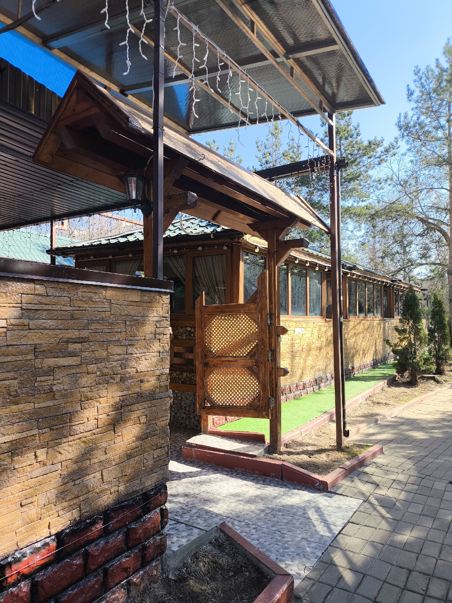 The image depicts a structure with a covered patio, likely a restaurant or cafe, located in what appears to be an outdoor area in Khimki, Russia. The structure features a combination of brick and wooden elements. A section of the exterior wall is made of a light brown, stone-like brick veneer over a red brick base. The patio covering is a corrugated metal or plastic material supported by metal poles. Wooden structures serve as entryways, adorned with decorative latticework. The area around the building features paving stones, grass, and trees. The lighting suggests it is daytime with bright sunshine. There is no indication of people or specific activities visible.