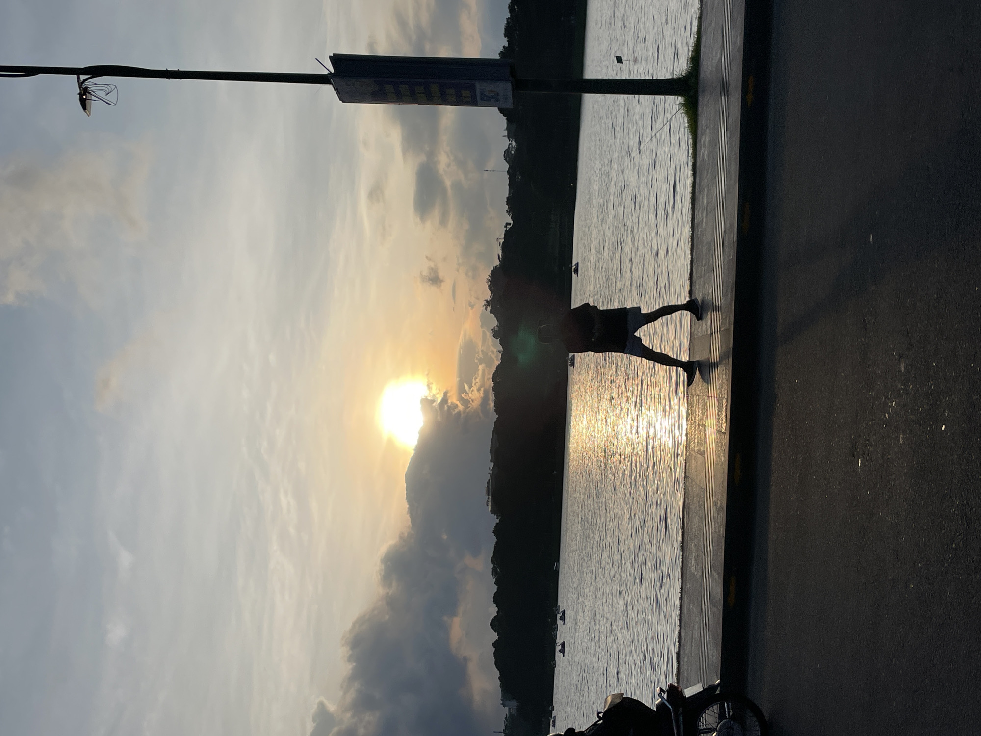 This image captures a sunset scene over Xuan Huong Lake in Da Lat, Vietnam. The sun, a bright golden orb, is low on the horizon in the upper left-center, partially obscured by dense, dark clouds. The sky transitions from a lighter gray-blue at the top to a warm orange-yellow near the sun, with scattered lighter clouds across the upper frame.
The mid-ground is dominated by the calm, rippled surface of Xuan Huong Lake, which reflects the golden sunlight in a shimmering path extending towards the foreground. Several white and dark pedal boats, some resembling swans, are visible on the lake, with small, indistinct figures inside them. Along the far shore, a dark silhouette of a tree line and distant hills or buildings stretches across the horizon.
In the foreground, a paved concrete walkway runs parallel to the lake's edge, featuring dark tactile paving strips. Adjacent to the walkway is a dark asphalt road. On the far left, the front wheel and engine area of a parked motorcycle are partially visible on the road or walkway.
A person, rendered in dark silhouette, walks from left to right on the walkway, wearing shorts and a t-shirt. Their long shadow is cast across the walkway and the road. On the far right, a tall, dark streetlamp post stands vertically, topped with a functional light fixture. Below the lamp, a rectangular signpost, also in silhouette, displays faint yellow markings. A thin fishing rod extends from near the base of the streetlamp towards the lake's edge. The overall scene is characterized by strong backlighting from the setting sun, creating prominent silhouettes and a warm, golden glow on the water.