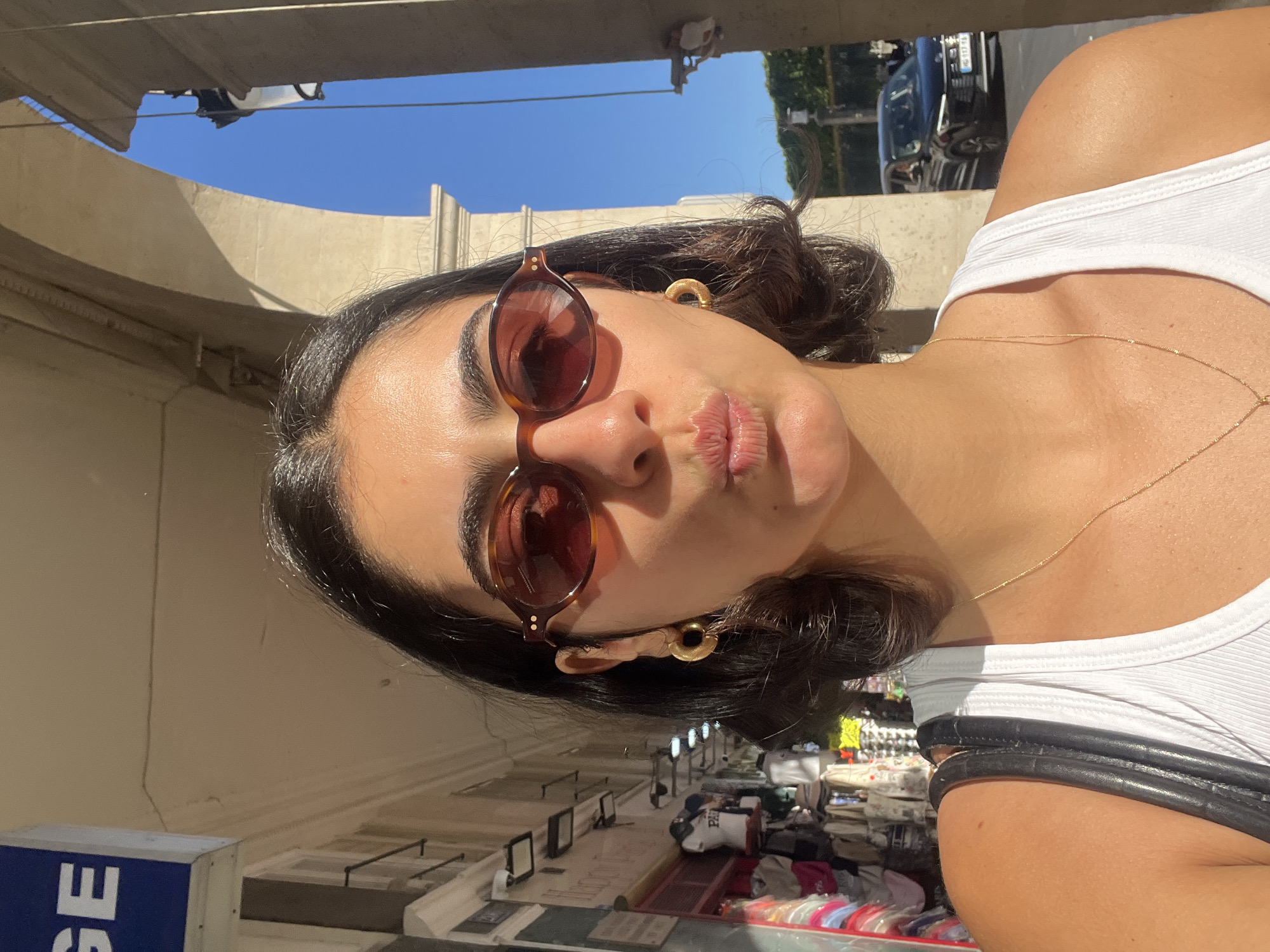 A close-up, dynamic selfie captures a young woman outdoors under bright, direct sunlight, likely in Paris, France, as indicated by the context. The image is rotated, showing her head tilted sharply upwards within the frame, creating an unconventional perspective.
The subject has dark hair, styled in a short bob, and is wearing round, tortoise-shell framed sunglasses that obscure most of her eyes. She sports gold hoop earrings and a delicate gold chain necklace, which rests on her bare skin above a white ribbed tank top. Her lips are pursed in a playful "duck face" or kissy pose.
The background reveals a clear blue sky and sun-drenched, light-colored buildings with distinctive curved architecture, casting strong shadows that suggest midday or early afternoon. Hints of an urban street scene are visible, including what appears to be market stalls with various items for sale and a dark-colored car in the upper right background. A black strap, possibly from a handbag, is slung over her shoulder.
Visible text includes the letters "SE" on a blue and white sign in the bottom left corner, part of an unknown word or shop name. The overall impression is one of a casual, sunny day spent exploring a lively city environment.