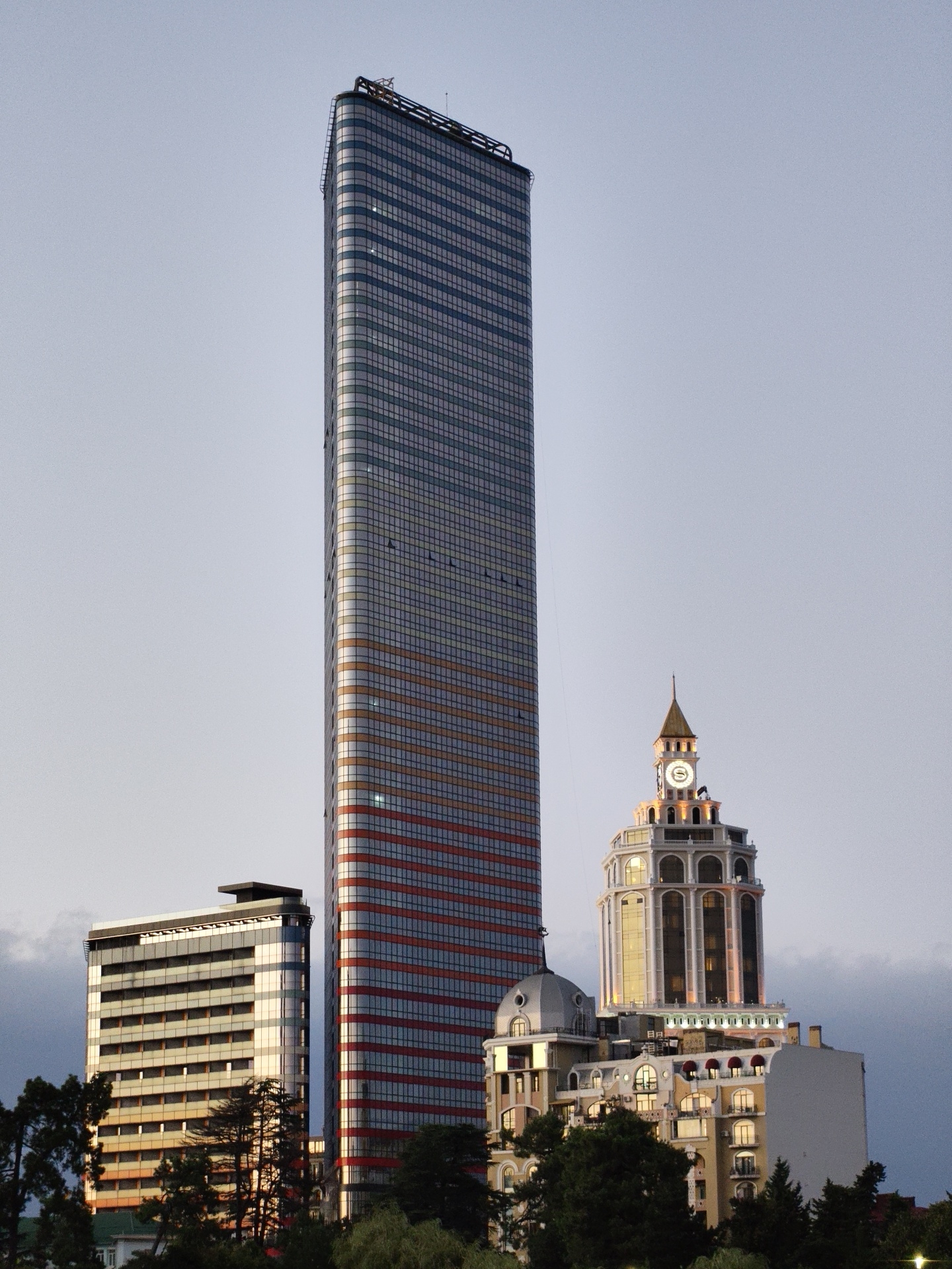 The image shows a city skyline at dusk, featuring several prominent buildings in Batumi, Georgia. The most striking structure is a very tall, slender skyscraper clad in glass and metallic panels, with horizontal bands of red and orange highlighting its facade. To the left of this skyscraper stands a shorter, rectangular building with a dark, flat roof and numerous horizontal windows. To the right, a more ornate building with a classical-inspired tower featuring a clock face and arched windows is illuminated. This tower is topped with a golden spire. Below the tower, a multi-story building with a pale facade and arched windows is visible, with some red awnings over some of the windows. Dark green trees are prominent in the foreground, obscuring the lower parts of the buildings. The sky above is a pale, overcast blue, indicating either early morning or late evening. The overall scene depicts a modern urban environment with a mix of architectural styles.