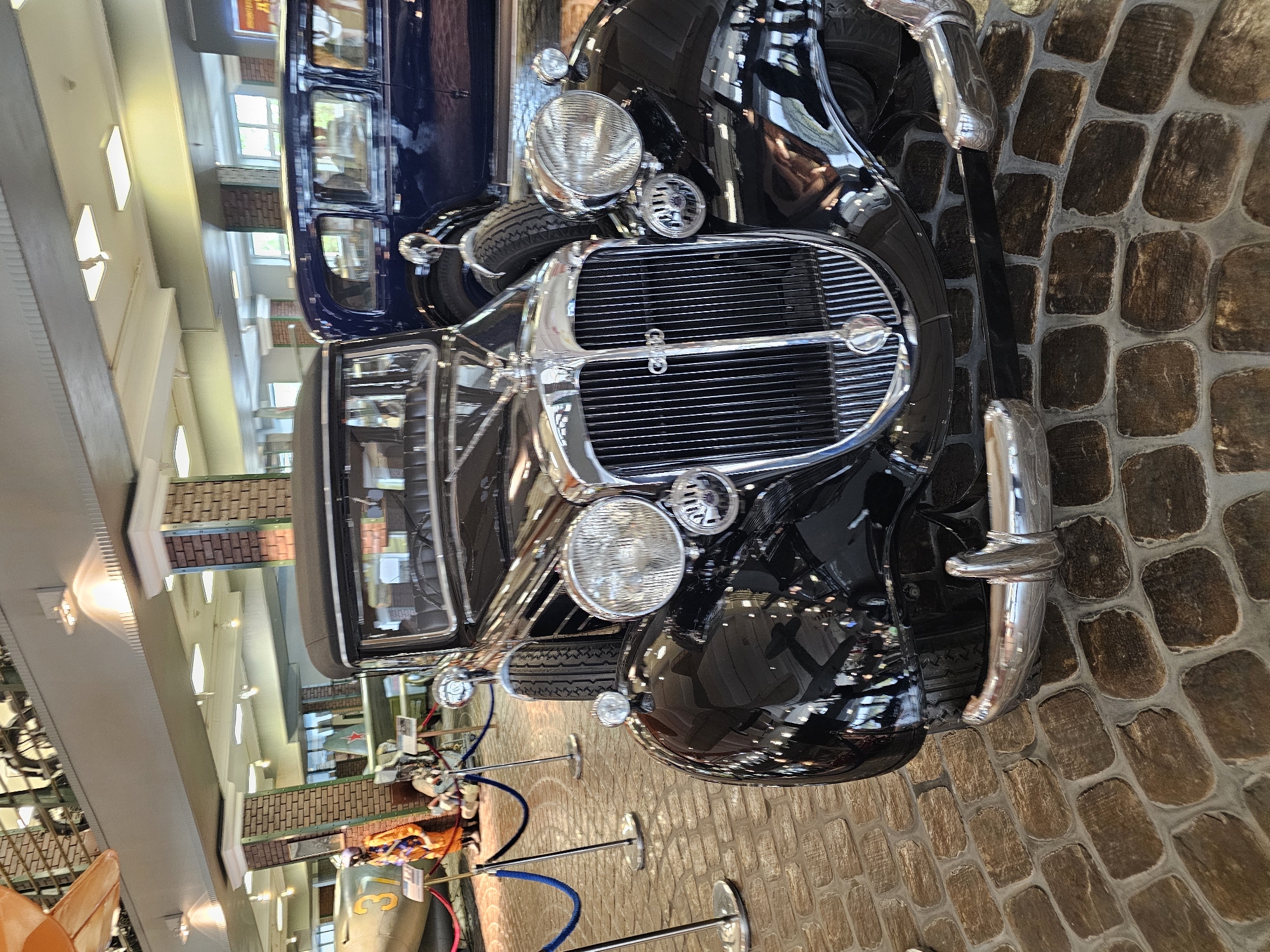 The image shows a black vintage car with a large, shiny chrome grill and headlights, situated inside a building with a brick floor. The building appears to be a museum or exhibit space. The car is near a roped-off area, likely to prevent visitors from touching it. In the background, there are other exhibits and historical vehicles. The lighting is bright, illuminating the car and other objects in the museum. The location is in the city of Архангельское, Russia.