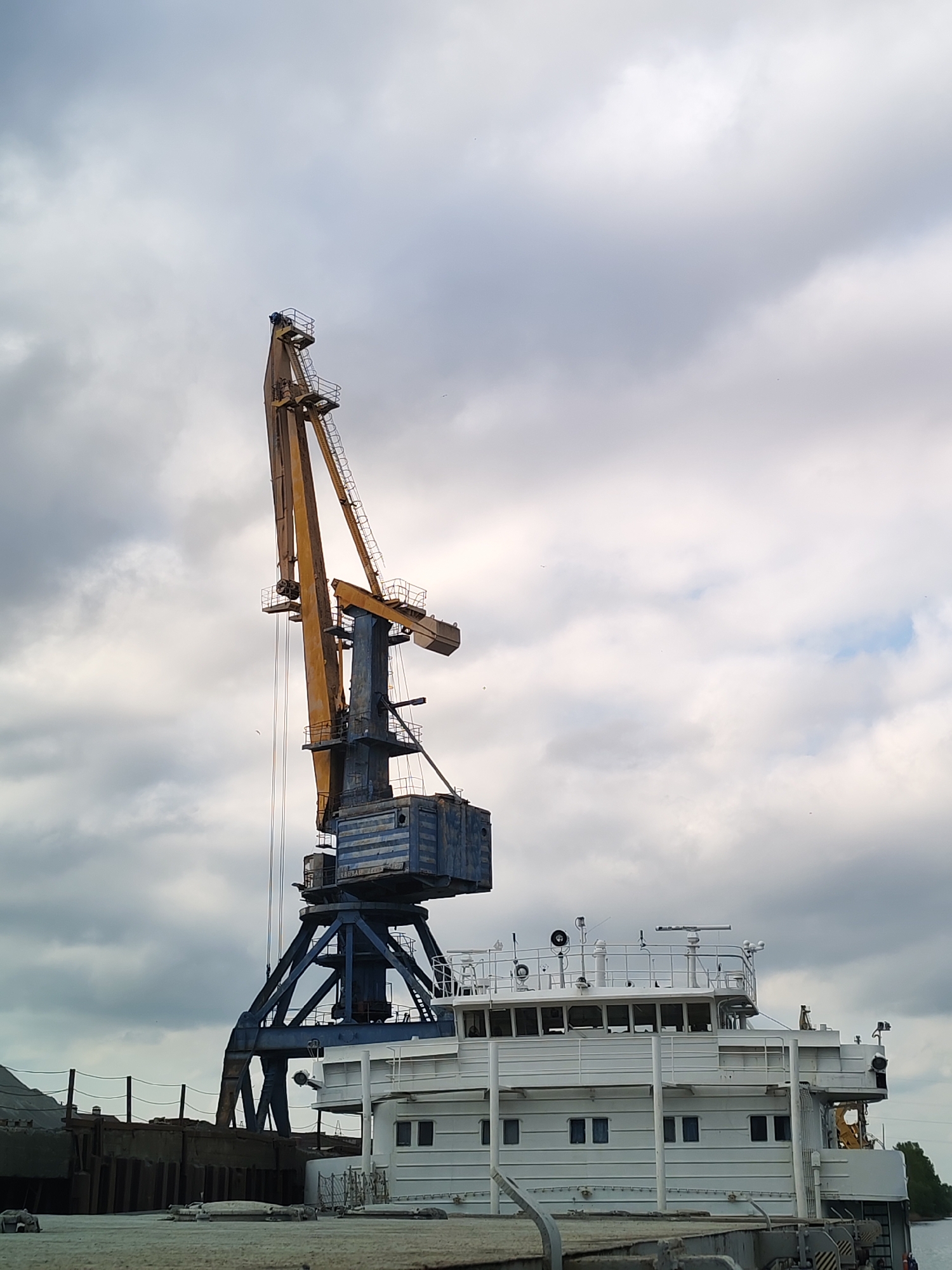 The image depicts a port scene in Tver, Russia, characterized by a large gantry crane positioned adjacent to a multi-tiered, white vessel (likely a barge or river boat). The crane is predominantly blue and yellow, with a long extending boom. The vessel features multiple windows and a top deck with railings and various navigational equipment. The background consists of a cloudy sky. Additional industrial structures and a strip of vegetation along the riverbank can be seen in the background. The foreground includes part of the dock.