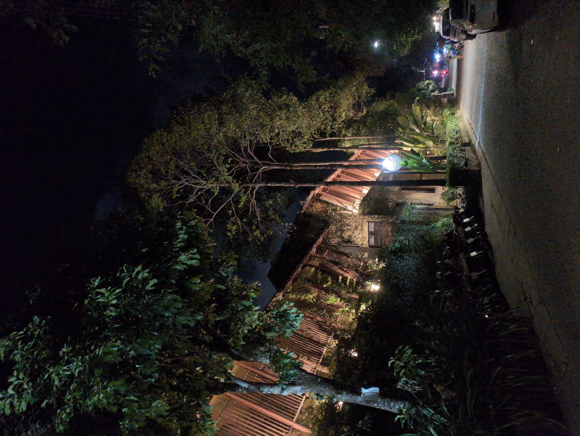 The image shows a nighttime scene in Phú Quốc, Vietnam, featuring a building with a prominent wooden lattice facade and a red tile roof. The building is partially obscured by trees and lush vegetation. A street runs alongside the building, with parked scooters visible. The scene is illuminated by a streetlight and the building's lighting, which casts a warm glow. Vehicles with illuminated headlights and taillights are present in the background.