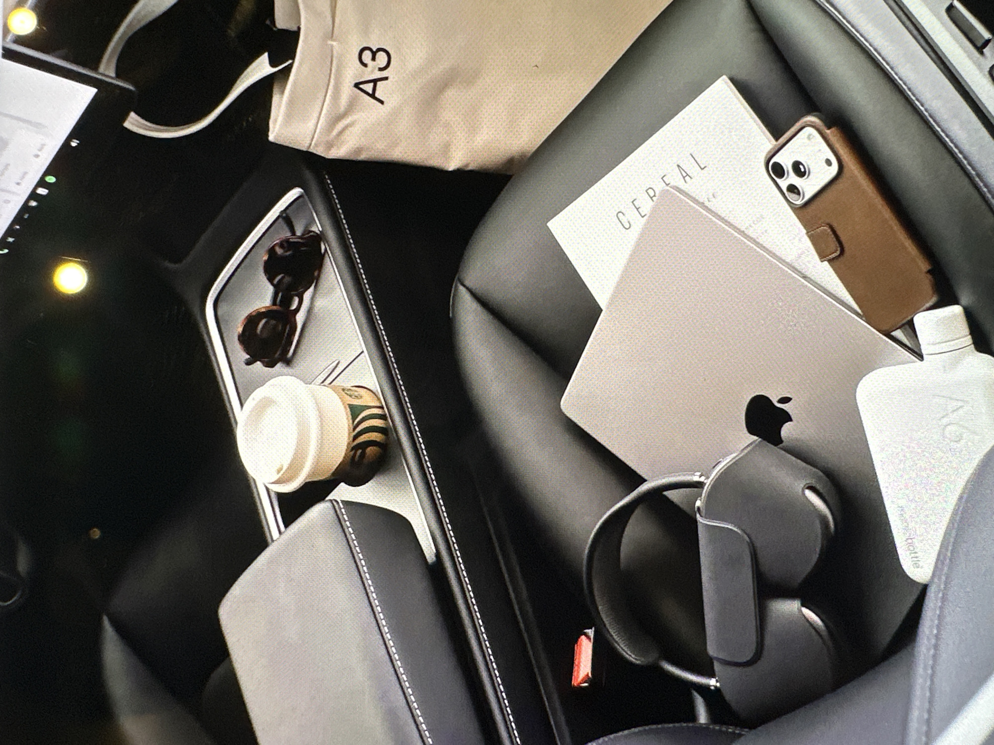 The scene is an interior shot of a vehicle, possibly a Tesla, laden with personal items. A gray MacBook with a black Apple logo is prominently placed on a black leather seat, adjacent to a white "CEREAL" book or magazine, and a brown leather cased iPhone, with a white A6 bottle nearby. A gray-black pair of headphones rests partially underneath the laptop. A beige "A3" tote bag lies near the top. On the console, there is a pair of tortoiseshell sunglasses and a takeaway coffee cup, labeled Starbucks. The location is not discernible from the image, therefore Yerevan, Armenia cannot be determined.