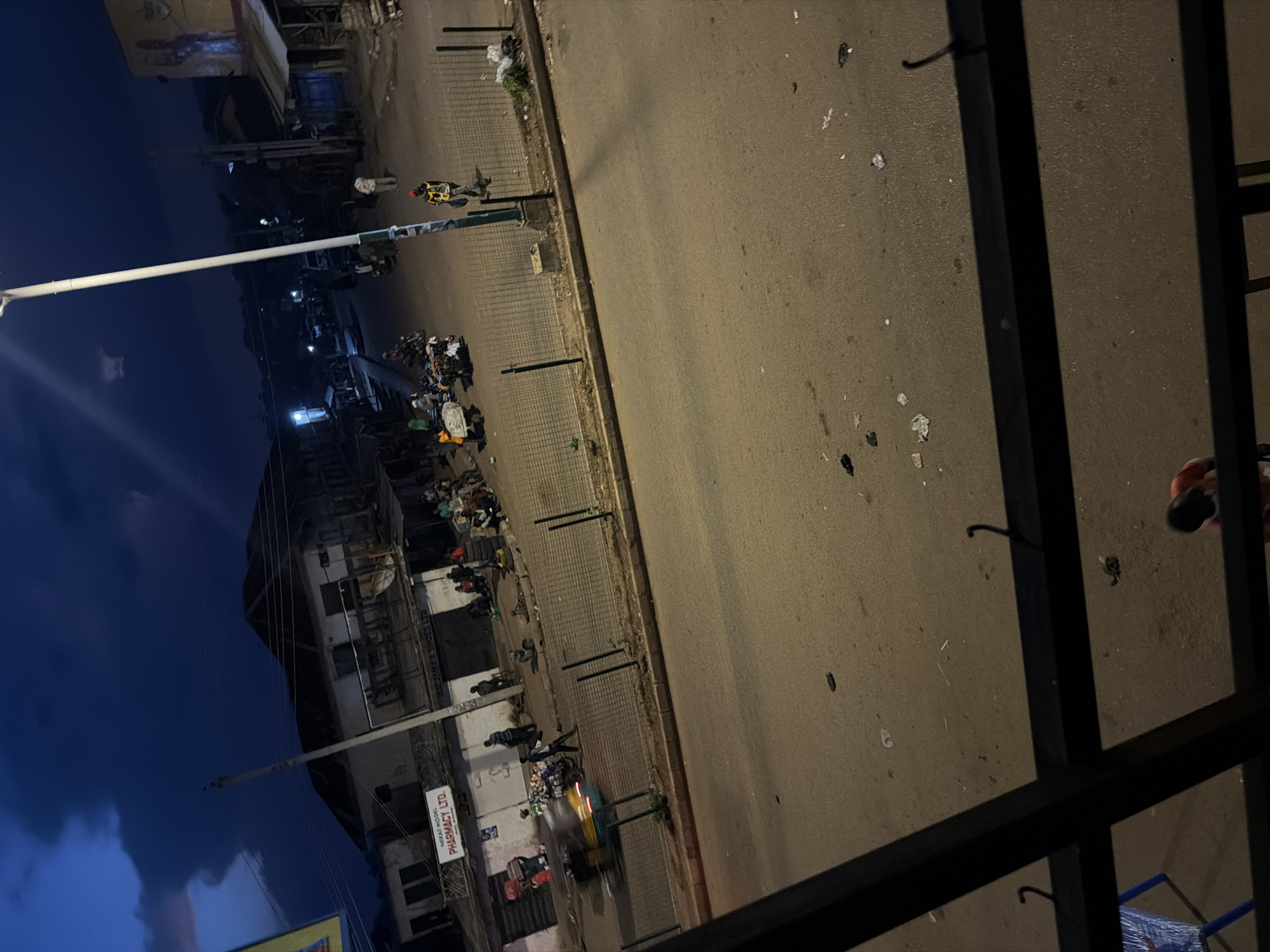 The image depicts a street scene at dusk in Bukuru, Nigeria, characterized by a mix of commercial buildings and a busy intersection. The sky is dark blue with some cloud cover. A prominent street lamp illuminates the area. On the left, a building with a sign that reads "PHARMACY LTD." is visible, with people gathered outside. Across the street, there are more storefronts and stalls. Several motorcycles and a motorized rickshaw are present, some in motion, suggesting active traffic. People are seen walking, standing, and interacting in various parts of the scene. The road is paved and appears somewhat worn, with scattered debris. A metal fence runs along the side of the road. In the foreground, the view is partially obstructed by a dark metal railing. The overall atmosphere is that of a lively urban environment transitioning into the night.