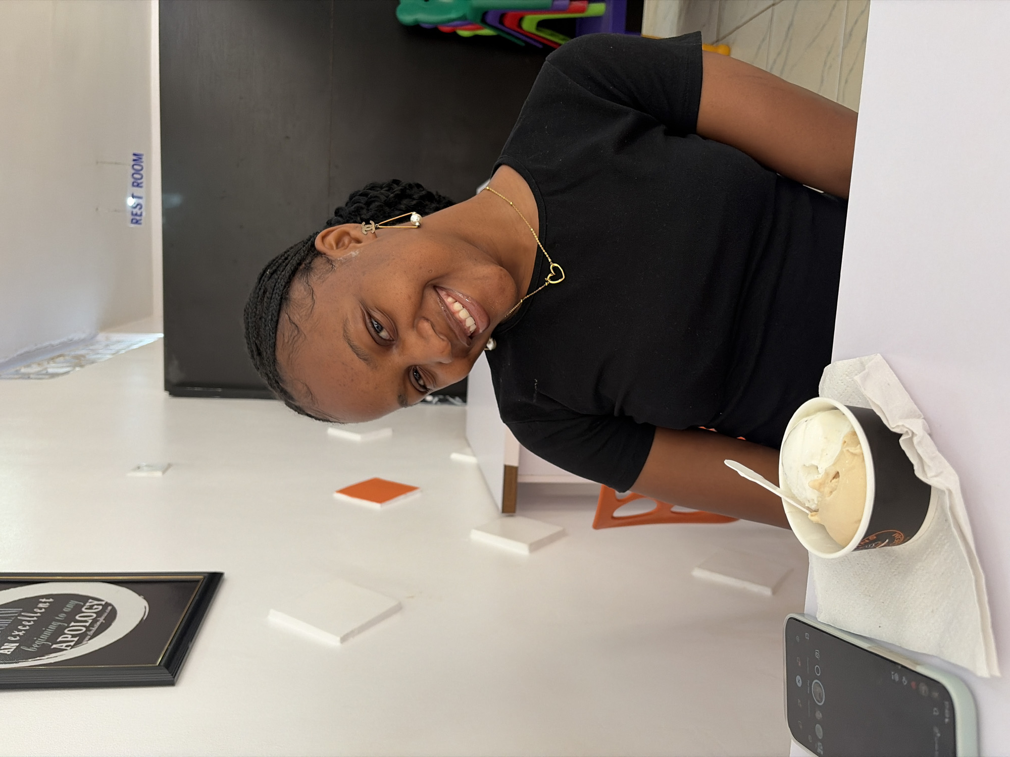 A medium shot captures a smiling young Black woman in an indoor setting, likely Bar Jirgi Summa, Nigeria, as provided by the context. She is wearing a black t-shirt, a gold heart pendant necklace, and distinctive gold and pearl drop earrings featuring a "CC" style logo. Her braided hair is styled neatly.
In the foreground to the right, a black-rimmed paper cup holds two scoops of ice cream (one light, one darker, possibly vanilla and caramel) with a white plastic spoon, resting on a white napkin beside a smartphone with a light blue/green case, its screen displaying media controls.
The background features a bright white floor with various small, square white and orange blocks scattered across it. A black-framed picture lies on the floor to the left, partially obscured, with visible text reading "an excellent beginning to any APOLOGY." A white wall with a blue sign indicating "REST ROOM" is visible in the upper left background. Behind the woman, glimpses of colorful plastic items (possibly chairs or toys) are visible. The woman is looking directly at the camera, conveying a cheerful and relaxed demeanor.