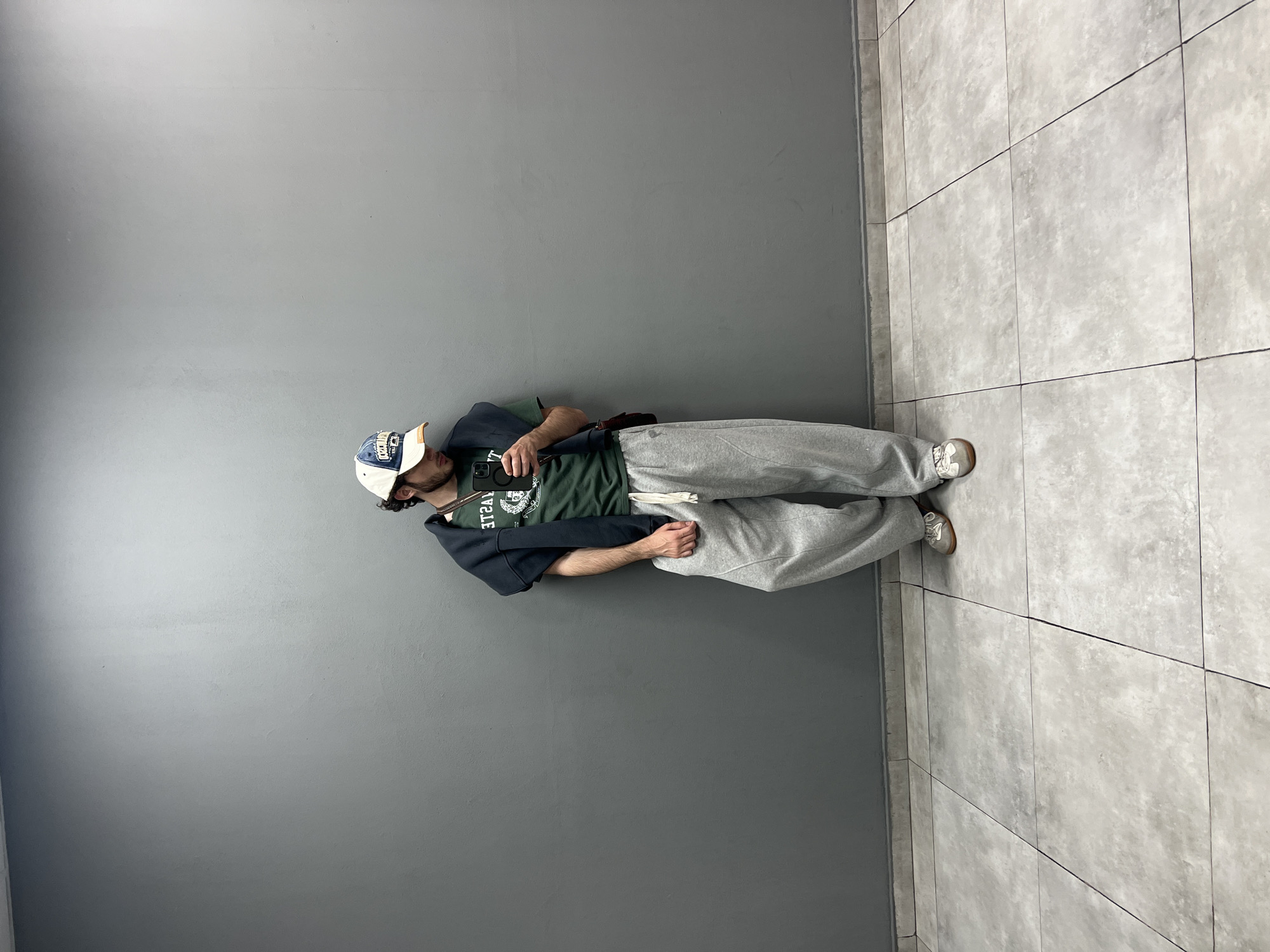 A man stands in front of a grey wall next to a tiled floor. The man is wearing a baseball cap, a dark green t-shirt with a white emblem, a dark colored hoodie, grey sweatpants, and tennis shoes. He is holding a cell phone. The location appears to be indoors, possibly a hallway. It is not possible to determine if this is in Yerevan, Armenia from the given image.