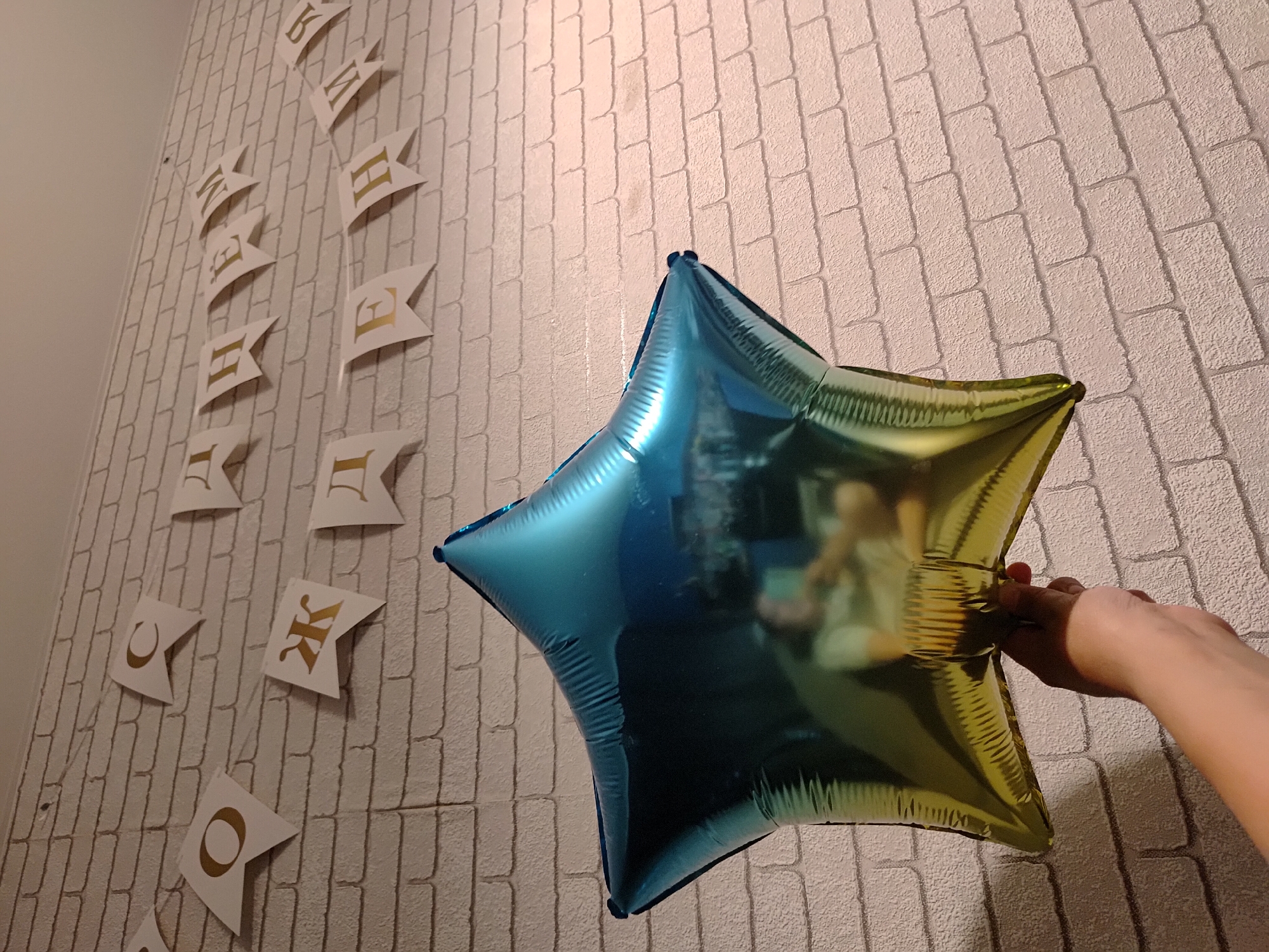 A person's hand holds a blue-and-yellow star-shaped balloon against a wall with a brick pattern. There is a banner with gold letters strung along the left side of the wall. The location is Марковский, Russia.