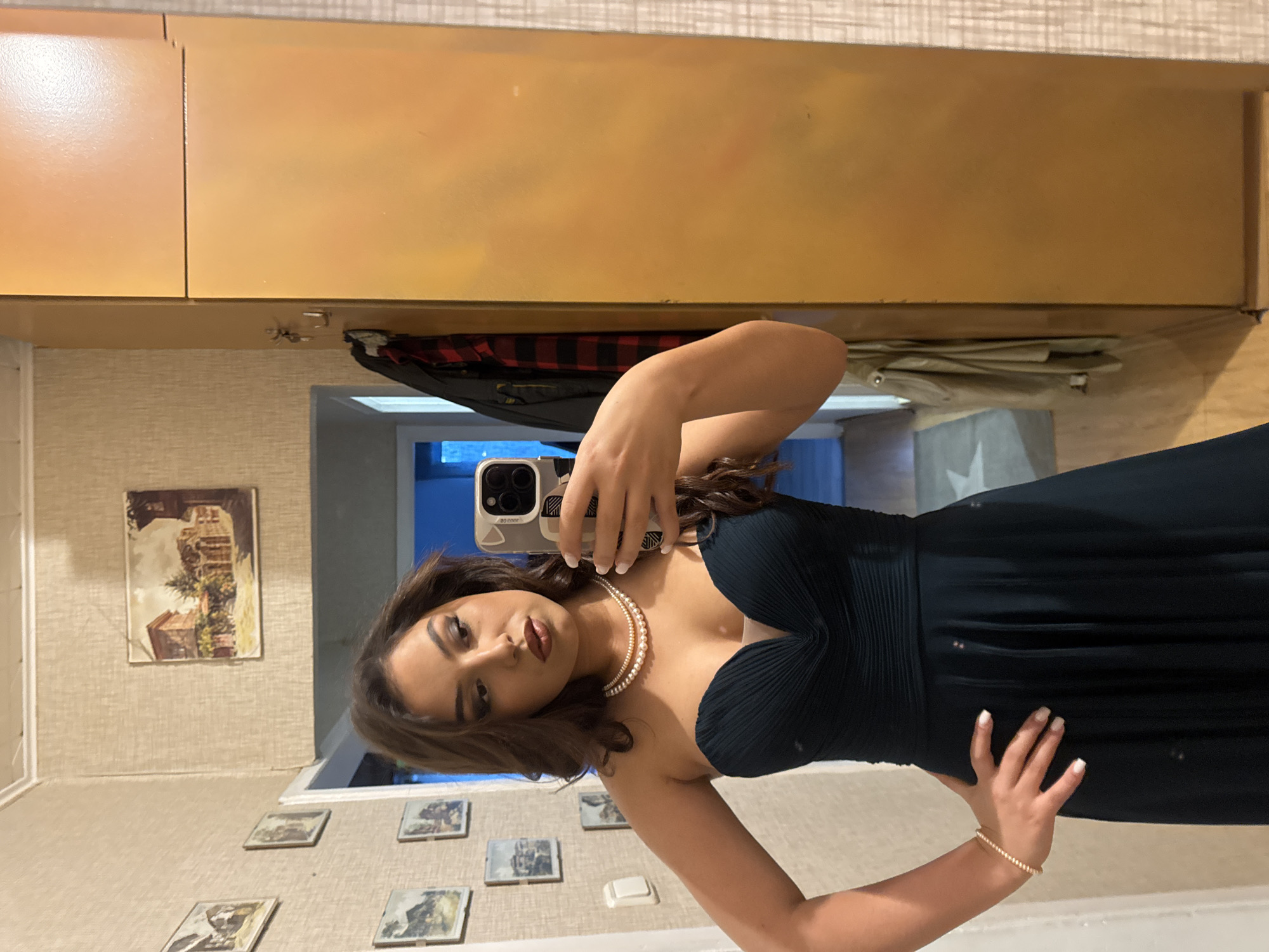 A young woman is standing indoors taking a selfie in front of a mirror. She is wearing a dark green, strapless formal dress, a pearl necklace, and a pearl bracelet. Her hair is brown and styled in waves. In her right hand, she is holding an iPhone with a protective case, using it to capture the image. Her left hand rests on her waist.
Behind her, the room appears to be a hallway or entrance area. The wall is covered in textured wallpaper, and there are several framed pictures hanging at various heights. Above, there are wooden cabinets, and a jacket is hanging from a hook. Through an open doorway, a glimpse of a room with a white star pattern on the floor is visible. The specific location in the city of Unknown, Unknown cannot be determined.