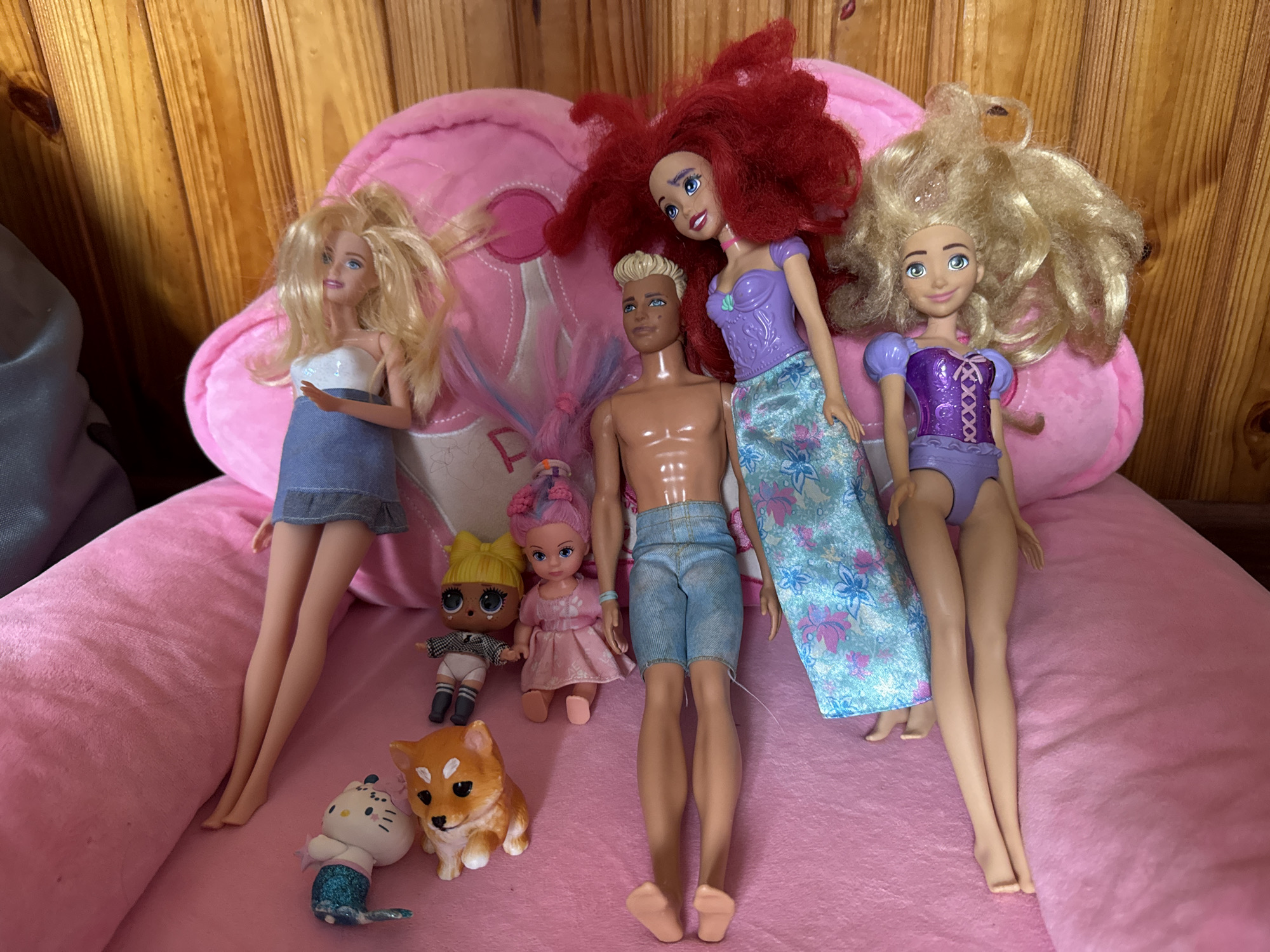 In a domestic setting, a collection of dolls is arranged on a pink plush surface, possibly a large stuffed animal, with a wooden planked background. The dolls include two Barbie-style dolls with blonde hair, one wearing a denim skirt and white top, the other in a purple swimsuit-like top and purple shorts. A male doll, resembling Ken, with blonde hair and a tanned complexion, is shirtless and wearing denim shorts. A doll with bright red hair and a purple top, reminiscent of Ariel from Disney's The Little Mermaid, is also present. Two smaller dolls are positioned near the larger ones: one with blonde pigtails and large eyes, dressed in a black and white checkered outfit, and another with pink hair and a pink dress. In the foreground, there are two animal figures: a white cat-shaped figure with a blue tail and a small orange dog with pointed ears, possibly a Shiba Inu. The lighting suggests an indoor environment.