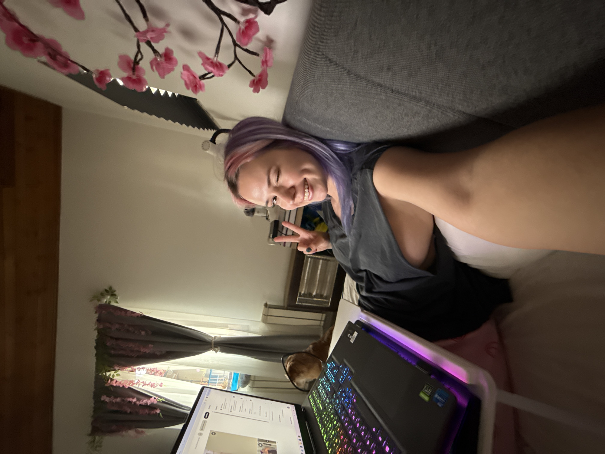 An indoor scene depicts a young adult female with pink and purple hair, wearing a dark grey t-shirt, reclining on a grey sofa or bed. She smiles and winks her left eye while making a peace sign with her right hand, suggesting a selfie. To her left, a black gaming laptop with an RGB backlit keyboard sits on a white portable stand, displaying a webpage. Partially visible behind the laptop is a light brown dog wearing a protective cone collar. The room features a window with grey and sheer white curtains, decorated with artificial pink wisteria and green foliage. Through the window, a blue balcony railing and other buildings are visible. A black decorative piece with pink cherry blossoms is mounted on the wall to the subject's right. Other visible objects include a white hairdryer, a radiator, a dark wooden cabinet, and a wooden ceiling beam. The precise location within the city of City of Zagreb, Croatia, cannot be determined from the image.
