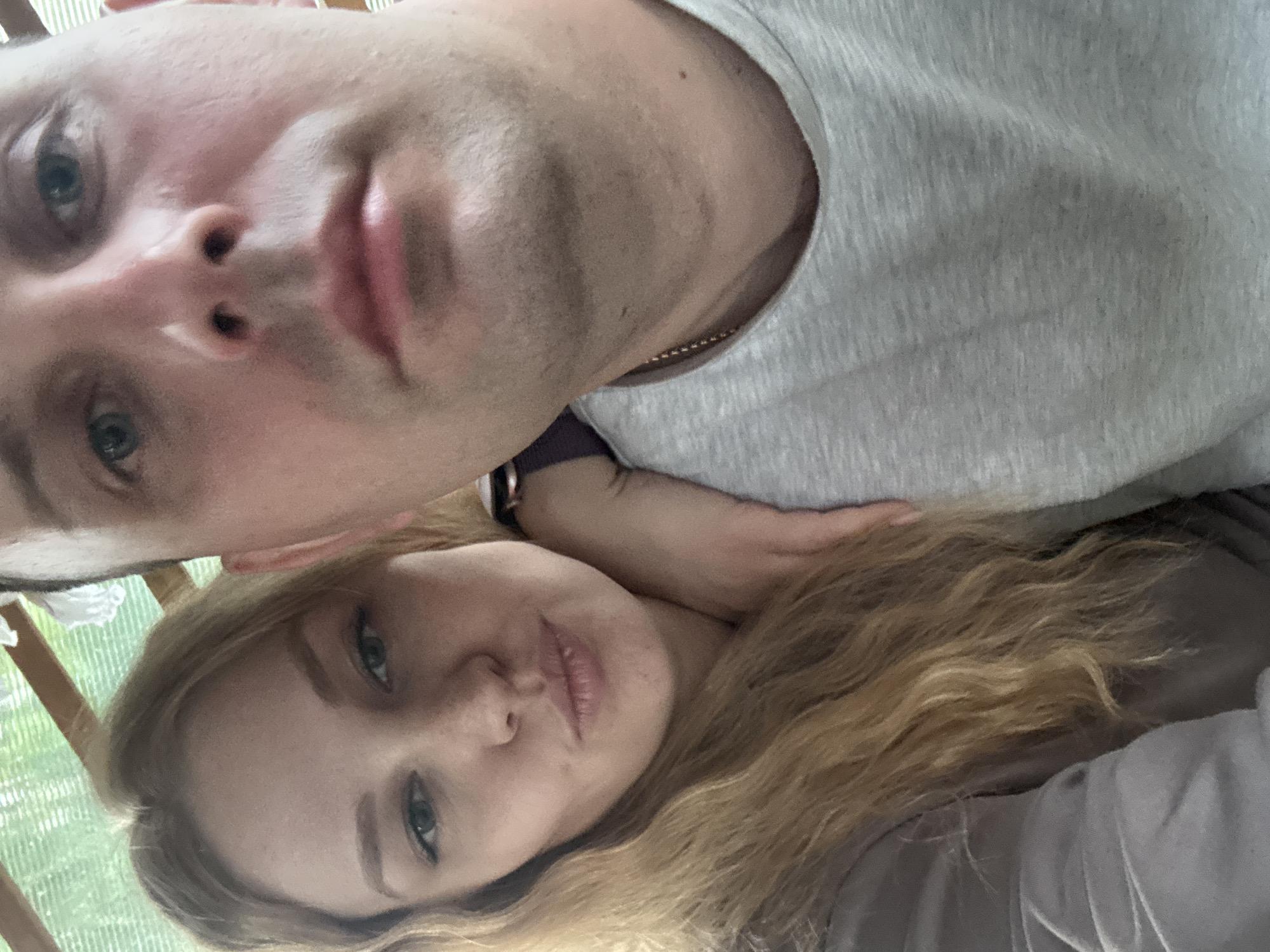 This image captures two individuals, a woman and a man, in a close-up, selfie-style photograph taken in Rozhdestveno, Russia.
The woman is positioned on the left side of the frame. She has long, wavy, blonde hair and light-toned skin. Her eyes are light-colored, and she has visible eyebrow definition and eyeliner. Her lips are pursed, and she looks directly at the camera. She is wearing a light brown or taupe long-sleeved garment. Her right arm is visible, resting on the man's shoulder or upper arm, indicating close physical proximity.
The man is positioned on the right, occupying approximately two-thirds of the vertical frame. He has short, dark hair, light-colored eyes (blue or grey), and visible stubble on his chin and jawline. He is wearing a light grey, possibly heathered, crew-neck t-shirt. A thin gold-colored chain is visible around his neck, resting above the t-shirt collar. His face is angled slightly, with his gaze directed generally forward.
The background is out of focus, indicating a shallow depth of field. Visible elements include a translucent or mesh-like overhead structure, possibly a canopy or greenhouse roof, with green vegetation discernible through it. A light-colored wooden beam runs across the upper-central background. The lighting appears to be natural daylight.