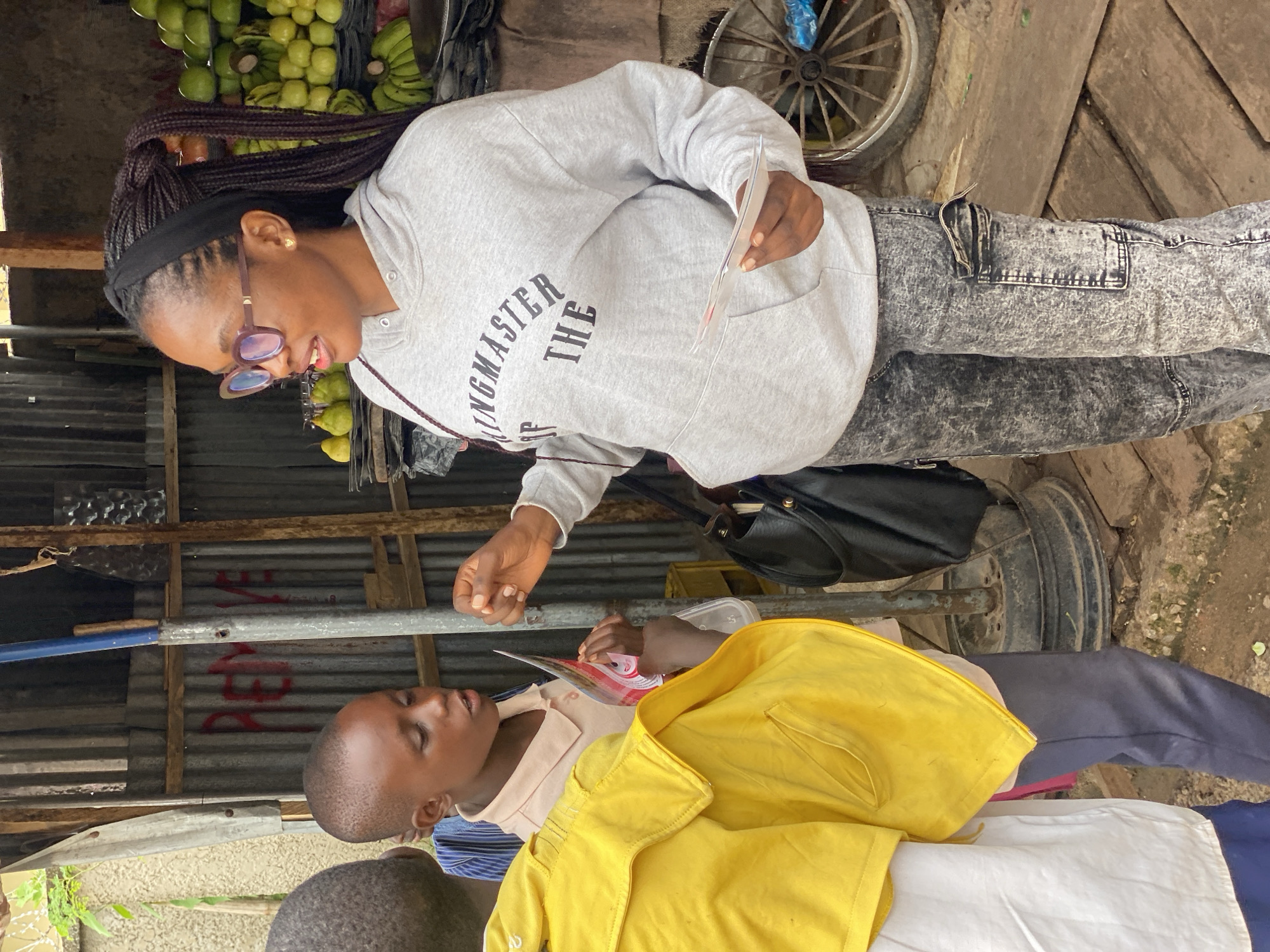 In Dadi Riba, Nigeria, a woman wearing a grey hooded sweatshirt with "KINGMASTER OF THE" printed on it and dark, speckled cargo pants stands on a dirt path. She is holding brochures and speaking to two young boys. The boy to her left wears a light pink collared shirt, and the boy to his left wears a bright yellow jacket over a white shirt. Behind them, a stall is constructed from corrugated metal sheets. Fruit, including green apples and bananas, is visible in the background, along with a wooden cart wheel. The setting appears to be an outdoor market or roadside stall.