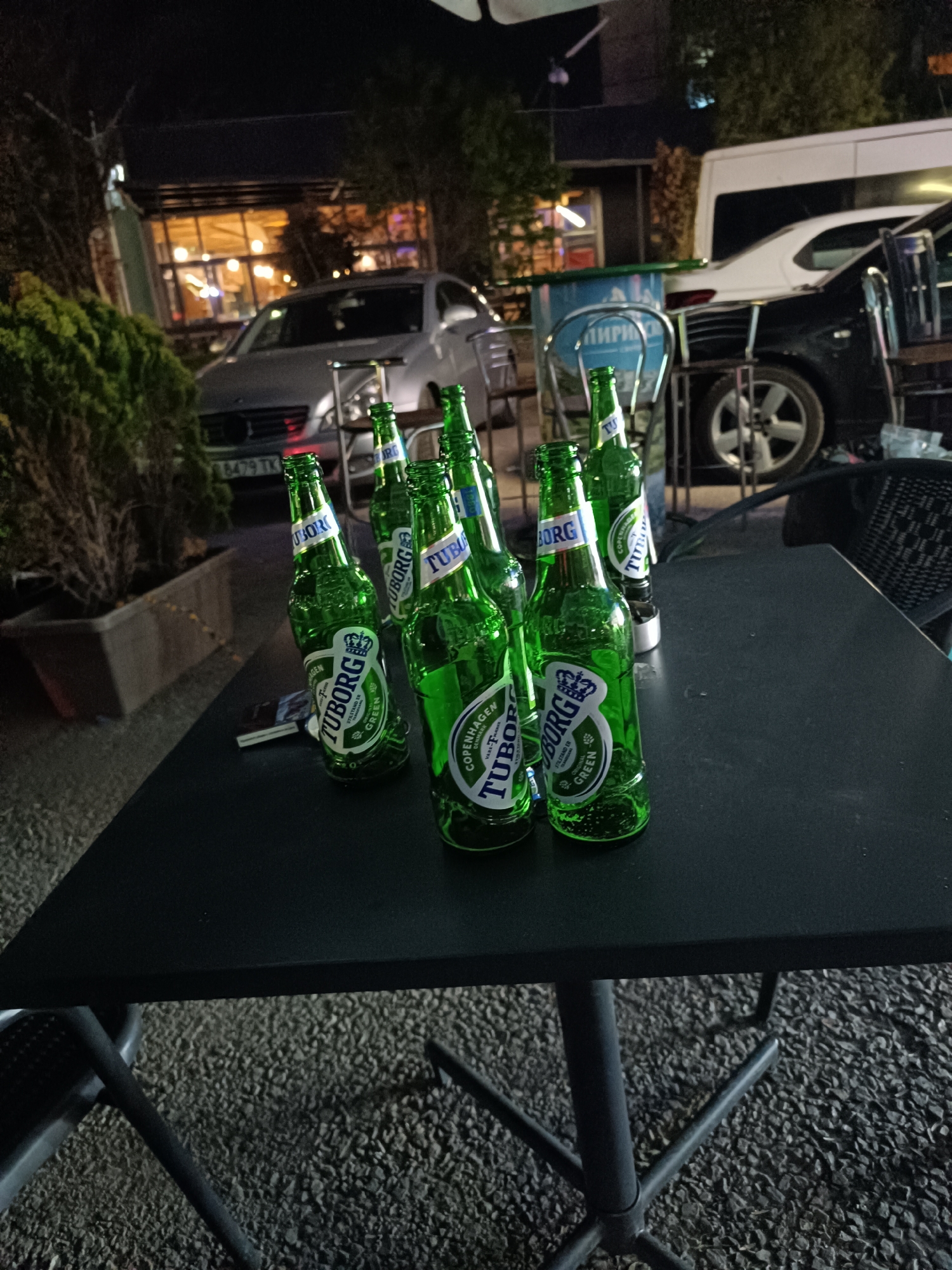 The image shows a table on the street outside, likely in Sofia, Bulgaria. Seven green Tuborg beer bottles are placed on the dark-colored table. The license plate of a Mercedes-Benz car reads "8479 TK". In the background, there's a bar or restaurant with bright lights, and behind the table, a cooler with the Cyrillic word "ЛИРИ" is visible. Other vehicles are parked nearby. The overall scene suggests an outdoor dining or drinking area at night. The ground has a gravelly texture.