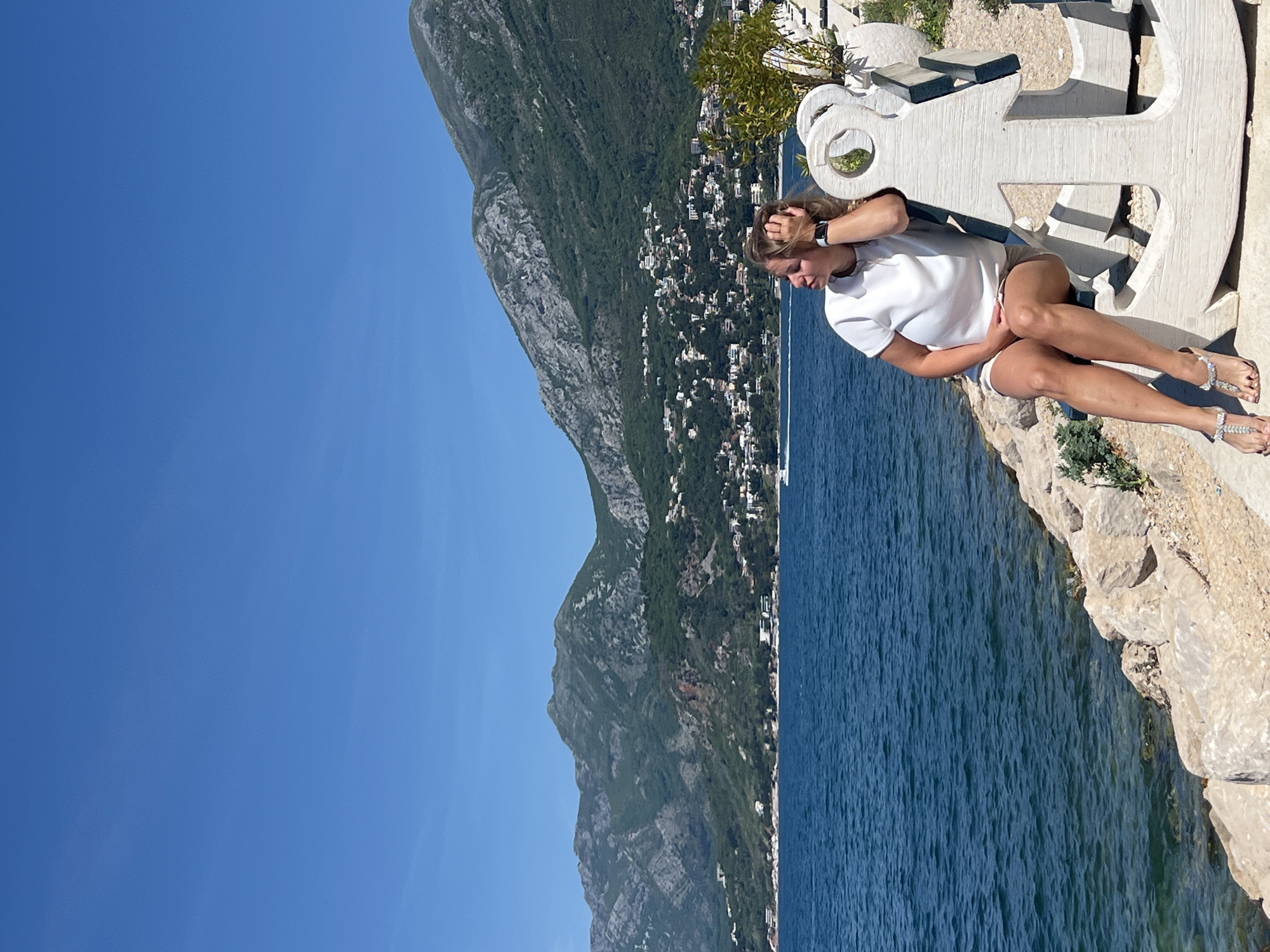 The image presents a scenic coastal view in Bar, Montenegro, captured on a bright, sunny day. A young woman with blonde hair is seated on a large, white, decorative anchor-shaped structure, which appears to be made of stone or concrete and serves as a viewpoint or seating area. She is wearing a white short-sleeved top, light-colored shorts, and sparkly sandals, with a watch visible on her left wrist. Her left hand is near her head as she looks down or to her side, seemingly in a relaxed or contemplative pose.
The anchor structure is situated on a rocky ledge overlooking a vast expanse of deep blue water, indicative of the Adriatic Sea. Across the water, a dramatic landscape of steep, green mountains rises directly from the coastline. Numerous white buildings are scattered along the foothills and lower slopes of these mountains, suggesting a town or village nestled along the coast. The sky above is a clear, vibrant blue, without any clouds. Strong sunlight is evident from the distinct shadows cast by the anchor and the woman. There is no visible text in the image.