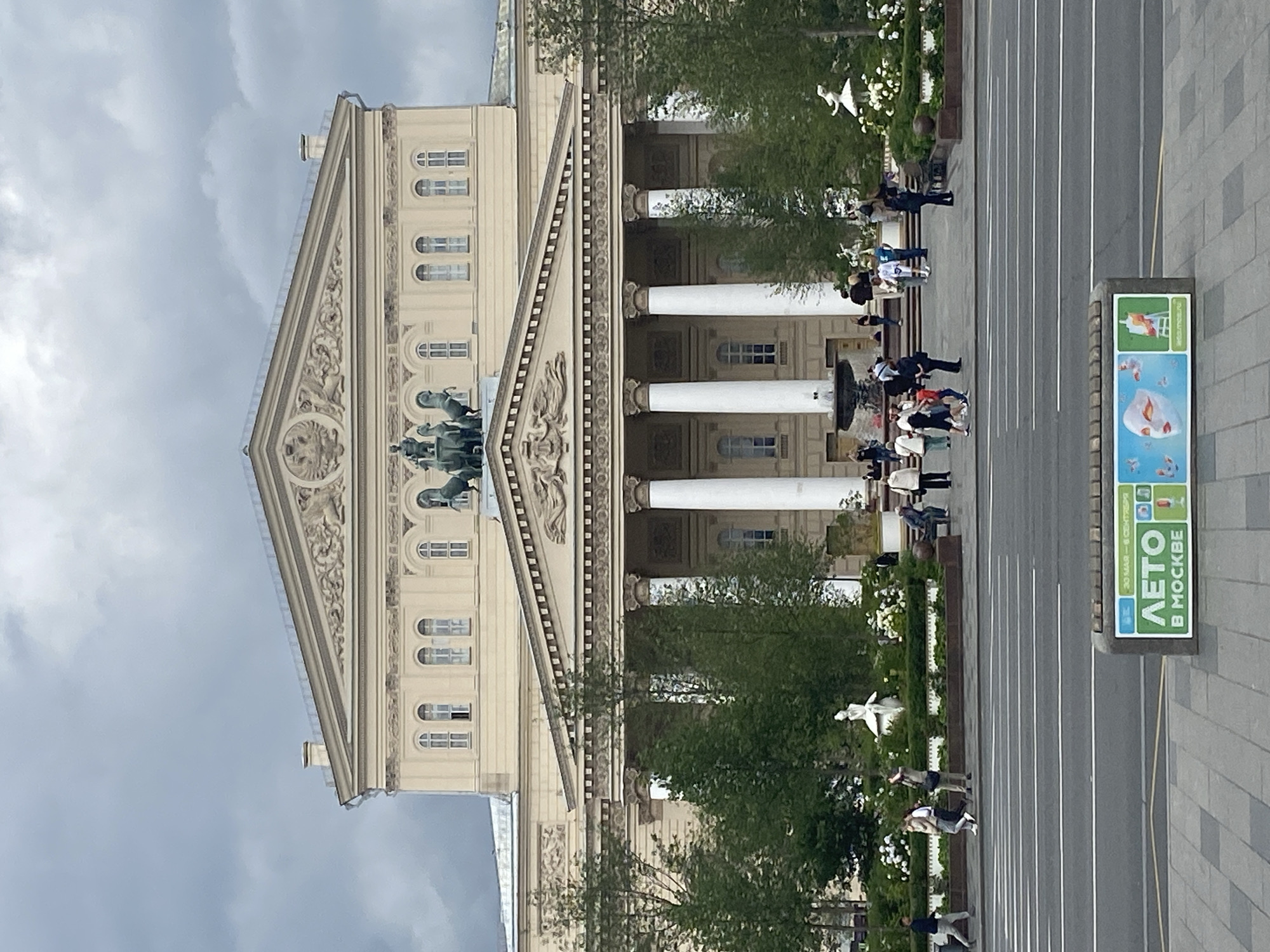 The image shows the Bolshoi Theatre in Moscow, Russia, under a cloudy sky. The neoclassical facade of the theatre dominates the upper half of the image, featuring a prominent portico with large white columns. Above the portico, a pediment is adorned with intricate relief sculptures, including a quadriga, a chariot drawn by four horses, at its apex. The building's beige exterior, with its arched windows and decorative detailing, is visible.
In front of the theatre, a public square or plaza is lined with trees and flower beds. Several people are seen walking across the plaza, some individually and some in small groups. Statues are visible within the landscaping, including a white statue of a ballerina. A fountain is also present in the middle ground.
In the foreground, a multi-lane road runs horizontally across the image, with white lane markings. On the sidewalk or median beside the road, a rectangular advertising stand is positioned. The stand displays graphics and text related to an event or season, featuring imagery of masks, butterflies, and art supplies, with Russian text that appears to say "ЛЕТО В МОСКВЕ" (Summer in Moscow). The overall scene depicts a public space in Moscow, with the grandeur of the Bolshoi Theatre as the central focus.