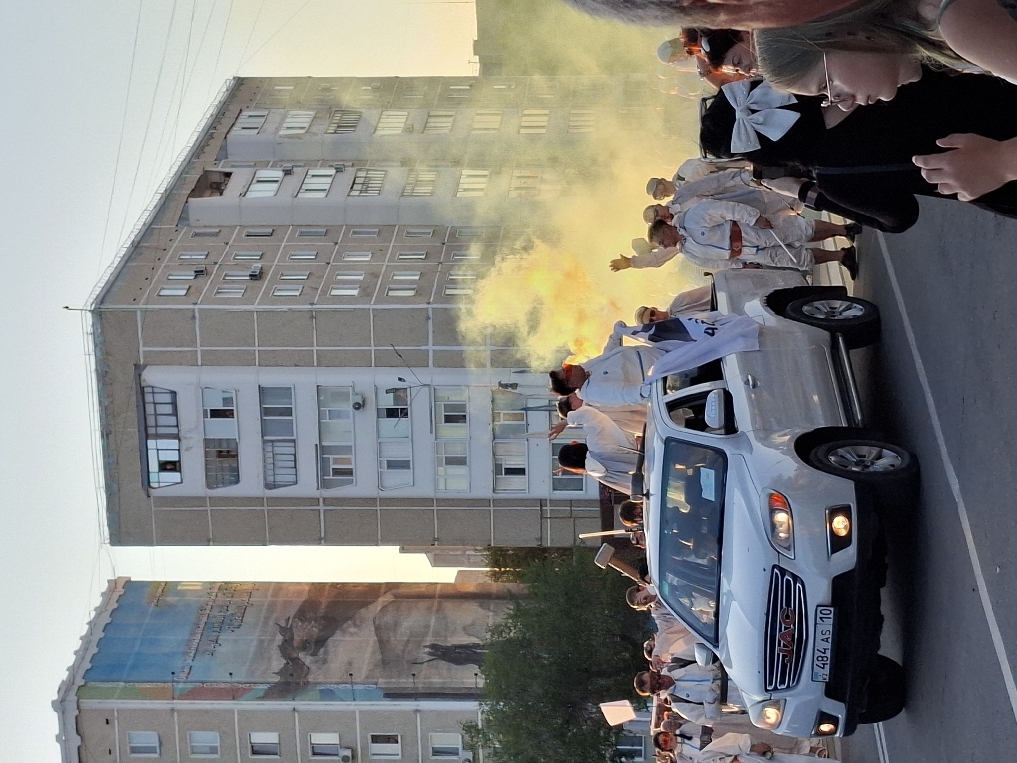 The image captures a street scene in a city, likely during a procession or event. A white JAC pickup truck is central to the composition, with several individuals dressed in white outfits standing on and around it. One person, wearing a red belt and goggles, stands on the truck's hood, holding a flag. Another person on the truck's side raises a hand. Yellow smoke is emanating from behind the truck, creating a hazy atmosphere.
In the background, two apartment buildings are visible. The building on the left features a large mural depicting what appears to be a dragon-like creature and text. The building on the right is a multi-story residential structure.
A crowd of people is gathered on the street, many also wearing white attire. Some individuals in the foreground are observing the procession. The scene is illuminated by natural light, suggesting it is daytime, possibly late afternoon given the warm tones. The license plate on the truck indicates it is from Kazakhstan.