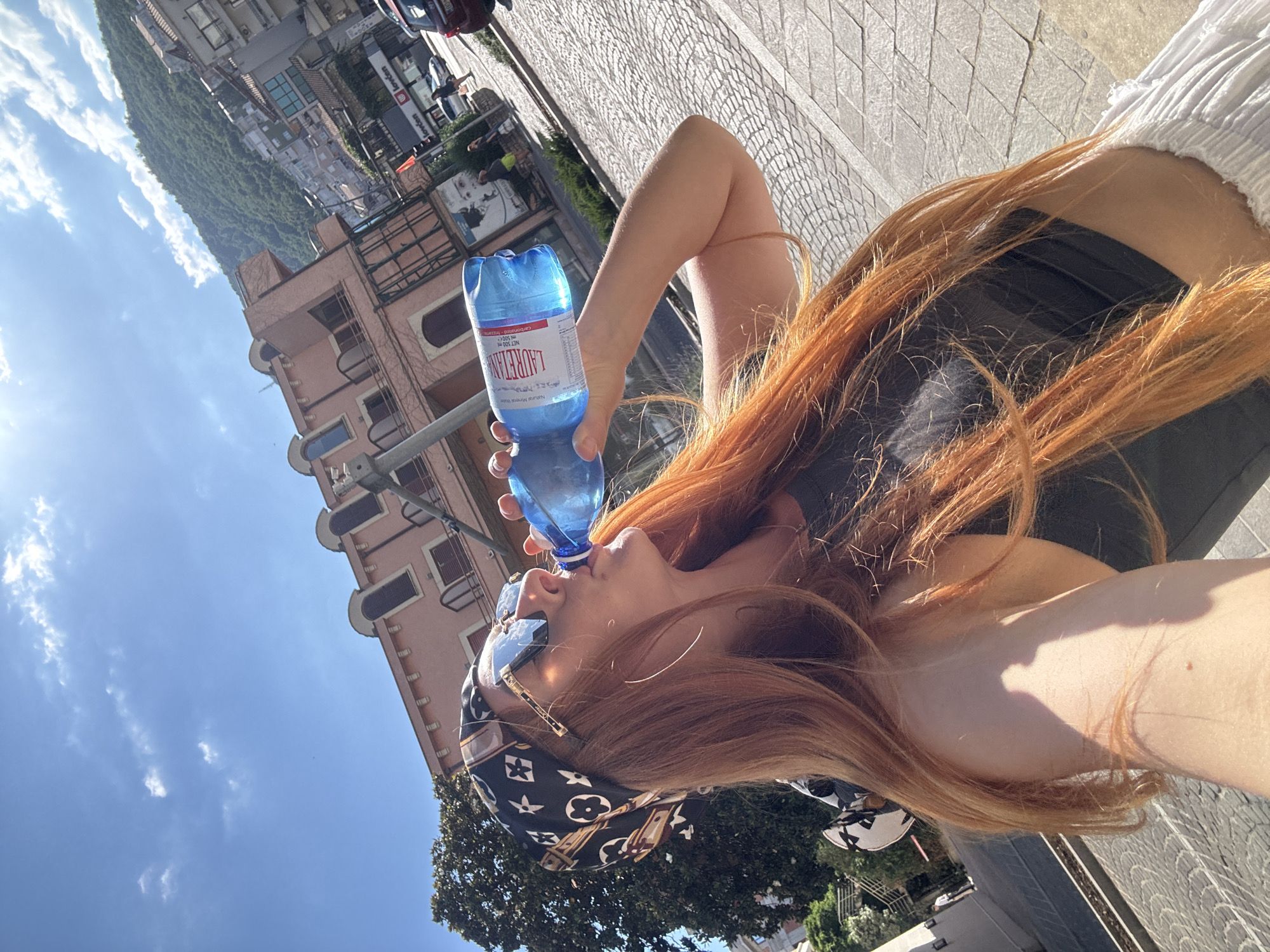 A woman with long, auburn hair, wearing sunglasses, a black bandana with a white pattern, and a black top, drinks from a blue plastic bottle of water labeled "LAURETAN." She is standing on a cobblestone street in Budva, Montenegro, with buildings and a tree in the background. The sky is blue with some clouds.