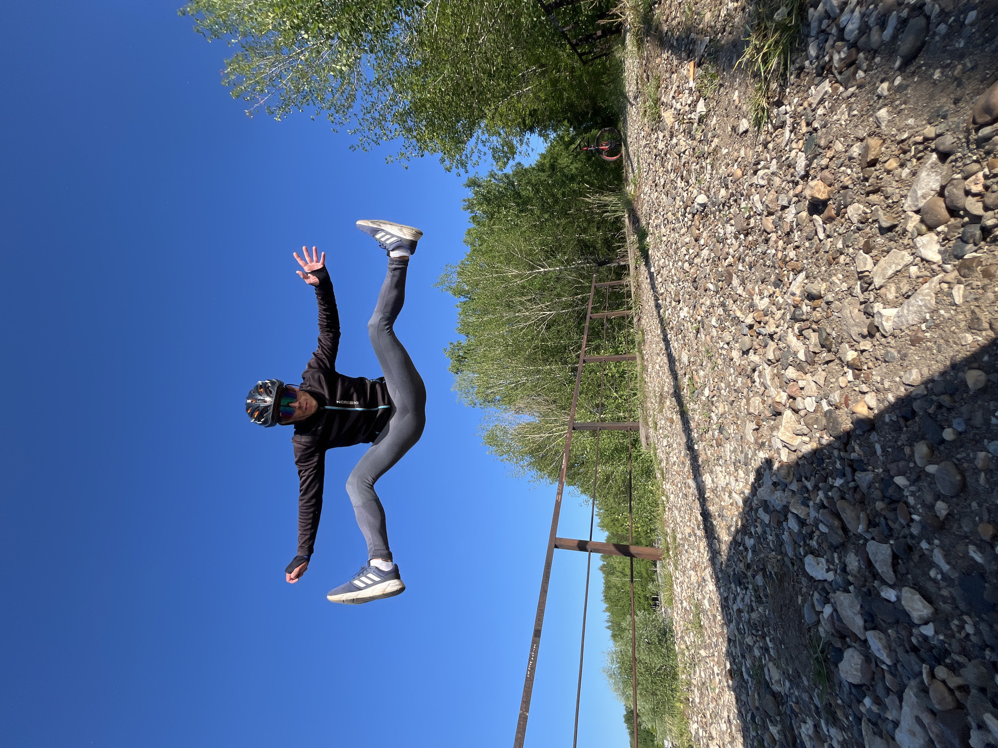 A person wearing a helmet, sunglasses, a black jacket, and gray leggings is captured mid-air in a wide jump, with legs extended horizontally to the sides. They are wearing Adidas sneakers. The scene appears to be outdoors on a sunny day with a clear blue sky. In the background, there is a brown metal fence with some graffiti on it, and beyond that, a line of green trees. A gravel path is visible in the foreground, leading towards the fence and trees. A bicycle with orange wheels is parked to the right in the background. The location is identified as Chaikovsky, Russia, specifically near a bridge or elevated walkway with a railing.